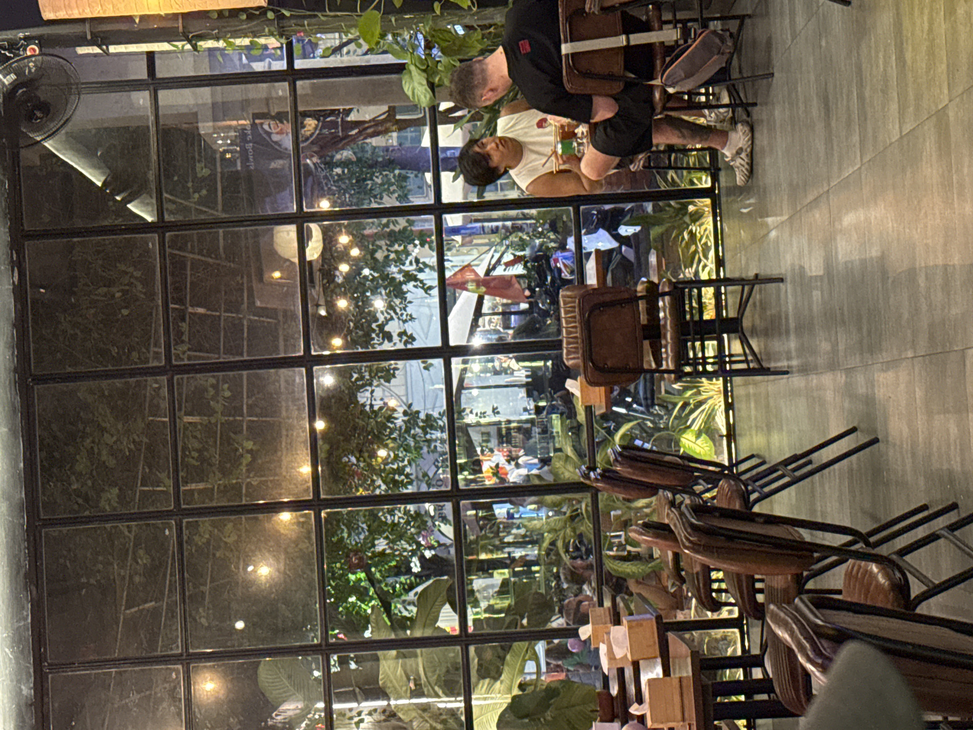 Captured from within a building through a large, multi-pane window is a scene from what appears to be a café or restaurant, situated in Đà Nẵng, Vietnam. Two men are seated at a table, engaged in conversation or an activity. One man wears a black shirt and shorts, while the other sports a white tank top. The interior of the establishment is decorated with abundant greenery, some of which is visible both inside and outside the window, creating a garden-like atmosphere.
Outside, the cityscape of Đà Nẵng is partially visible, with trees, buildings, and streetlights suggesting a bustling urban environment. A Vietnamese flag is prominently displayed in the background, confirming the location. Rows of empty chairs and tables are also visible inside. A ceiling fan is also visible.
