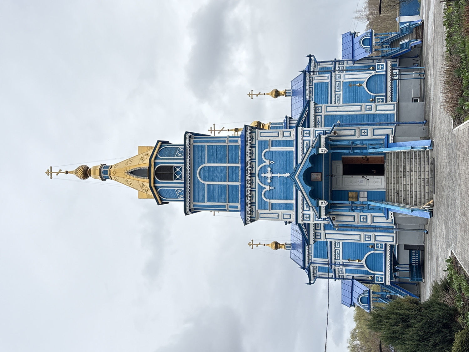 This image depicts an ornate wooden Orthodox church, likely located in Ulyanovsk, Russia. The church is distinguished by its vibrant blue and white facade, featuring intricate carved wooden detailing, archways, and decorative patterns. Multiple small crosses are integrated into the building's design.
The main structure includes a prominent entrance accessible via a set of stairs, leading to double doors. A striking tall bell tower or spire rises on the left, crowned with a large, multi-faceted golden onion dome and a slender golden Orthodox cross at its apex. Several smaller golden onion domes, each also topped with a cross, are visible on other sections of the church's roofline, contributing to its distinctive appearance.
The scene is set outdoors on an overcast, cloudy day, with a muted sky serving as the backdrop. A paved path runs alongside the building, and some green bushes and trees are visible on the right side of the frame. No people are present, and no specific activity or event is discernible. Near the main entrance, a small plaque with unreadable Cyrillic text is affixed to the wall. The church's bright colors and golden domes stand out against the subdued sky.