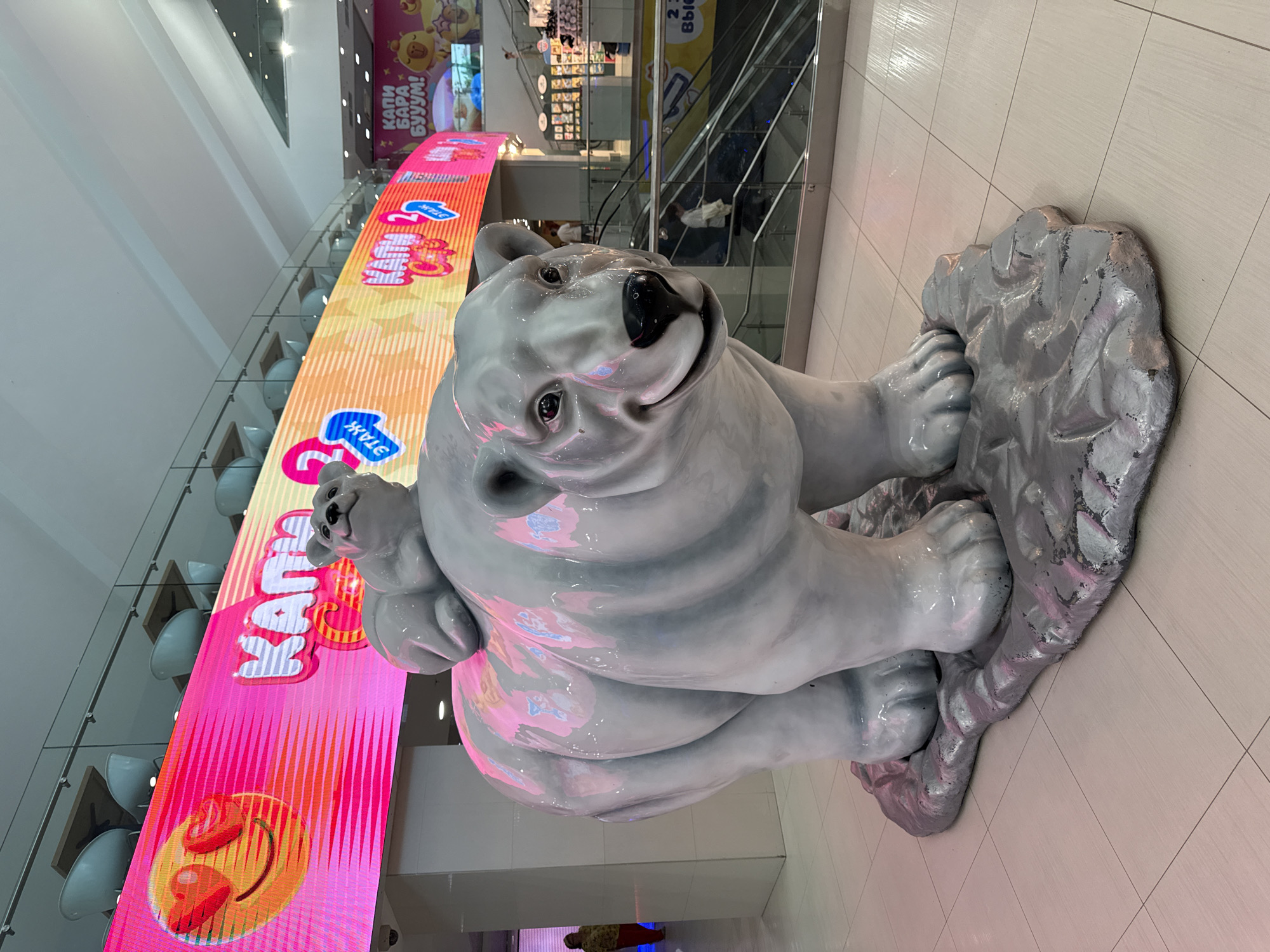 The image depicts a glossy, white-painted statue of a polar bear with a cub on its back. The statue is positioned on a grey, rock-like base. Behind the statue, there is a large digital display screen showing a bright pink and orange advertisement with text written in Cyrillic. An escalator can be seen in the background. The location is likely a shopping mall in Moscow, Russia. There are people partially visible in the background. The floor is tiled. The lighting appears to be artificial and bright.