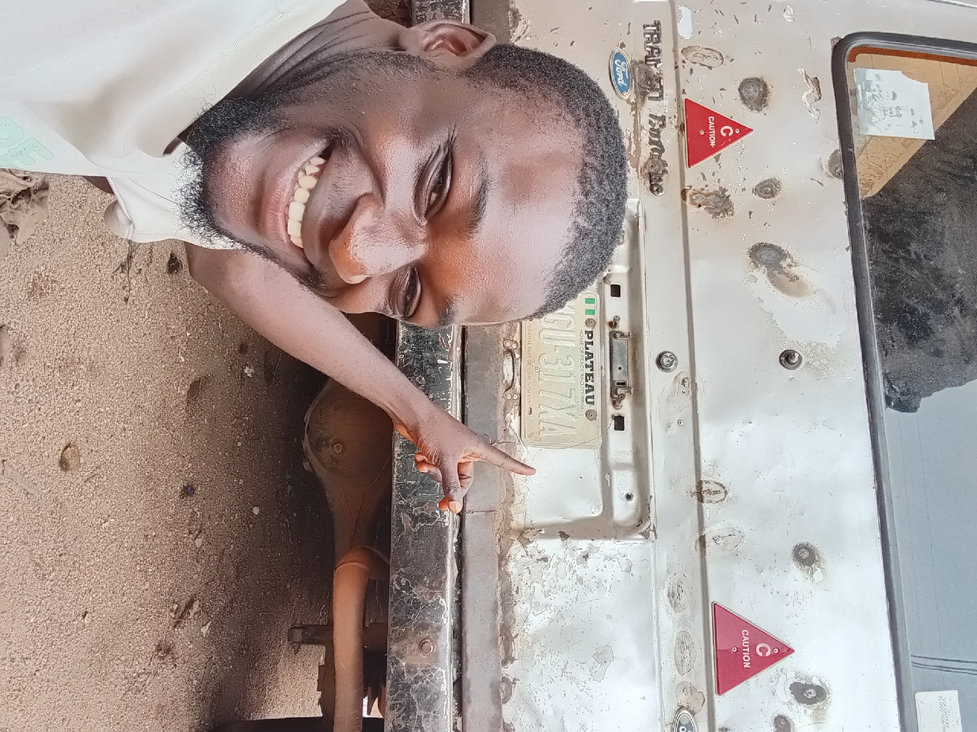 A young Black man is smiling and pointing at the rear of a beige Ford Transit van. The van has a license plate reading "PLATEAU GU 317XA" and two red triangular "CAUTION" stickers. The man is wearing a light-colored t-shirt. The background is dusty ground. The scene is likely outdoors in Bukuru, Nigeria.