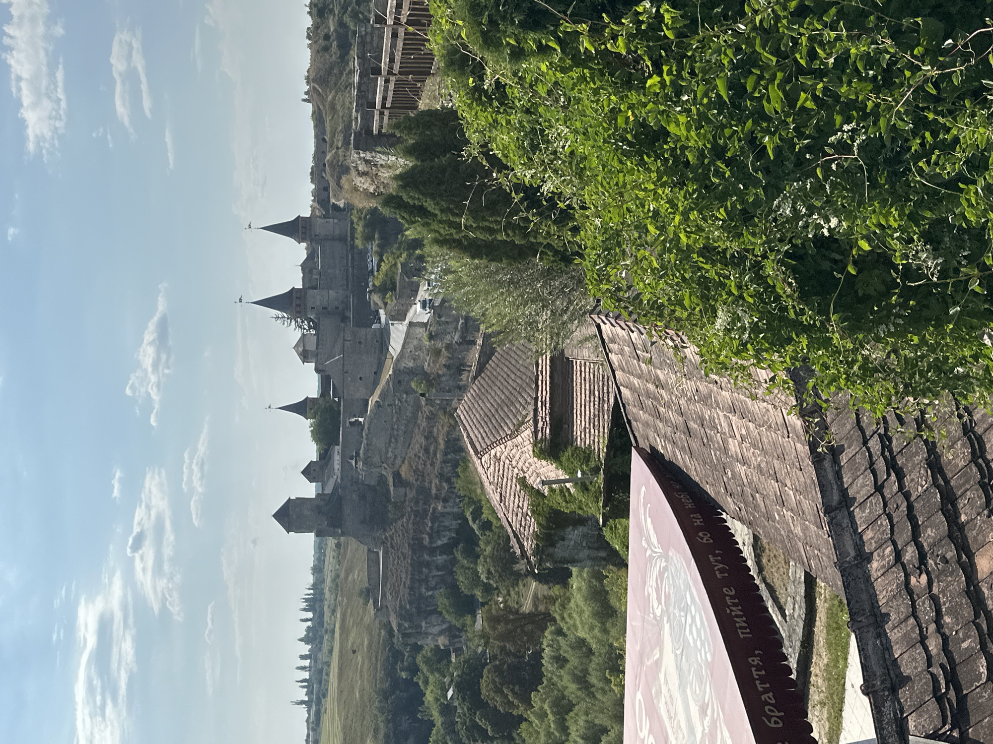 The image shows a panoramic view of the Kamianets-Podilskyi Castle in Ukraine. The castle, an ancient fortress, is situated on a rocky promontory, with its stone walls and conical towers dominating the skyline. The castle is surrounded by lush greenery, including trees and bushes, and a deep gorge. In the foreground, there are several tiled roofs of buildings, suggesting a settlement near the castle. A large red awning with text in Cyrillic script is also visible in the lower left corner. The sky is partly cloudy, with the sun casting shadows, indicating it is daytime. The overall scene depicts a historic landmark in a natural setting.