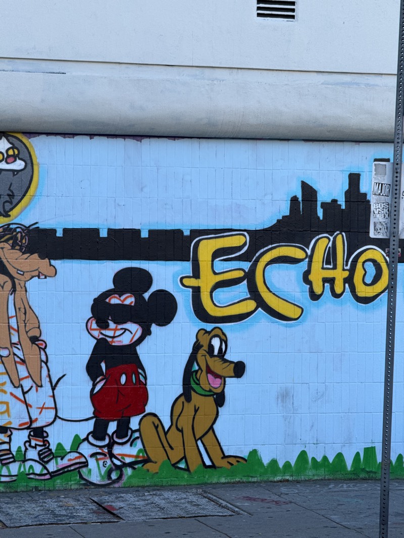 The image captures a mural painted on a light blue tiled wall, depicting cartoon characters. From left to right, there is Goofy, Mickey Mouse, and Pluto. Above them is the word “ECHO” written in yellow bubble letters with a black outline, positioned over a black silhouette of a cityscape. The scene suggests an urban environment, possibly Los Angeles, California, given the style of street art and the presence of a utility pole with posted signs.