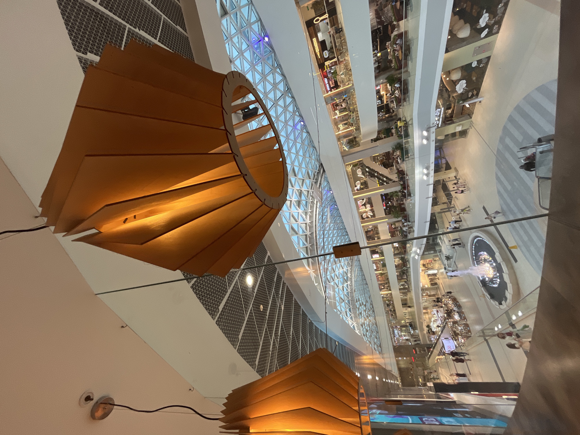 The image shows an interior view of a multi-level shopping mall. Prominent wooden lampshades hang from the ceiling, casting a warm light. The architecture features a modern design with a glass-paneled, geodesic-like ceiling that allows natural light to filter in. Below, several levels of shops and walkways are visible. People are seen walking, browsing, and sitting at cafes. In the center of the lower level, a fountain is active, with water spraying upwards. The overall impression is of a bustling and contemporary commercial space. This scene is located in the city of Sochi, Russia, likely within the "Morskaya Vola" (Sea Wave) shopping and entertainment complex, known for its modern design and amenities.