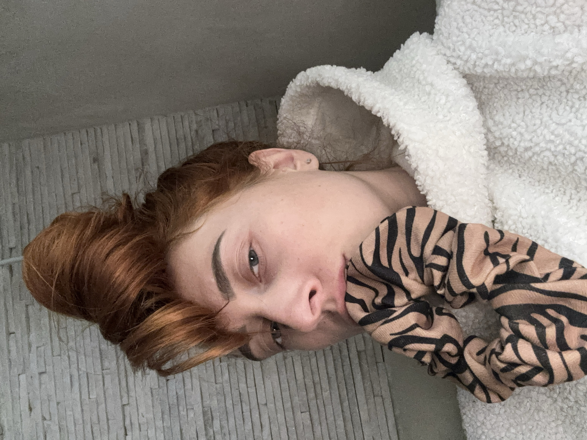 A young woman with auburn hair is lying down, likely indoors, as evidenced by the wall covering, a fuzzy white hooded robe, and a scrunchie with a zebra print near her face. Her face is slightly tilted towards the camera. The context of Budva, Montenegro is not apparent from the image.