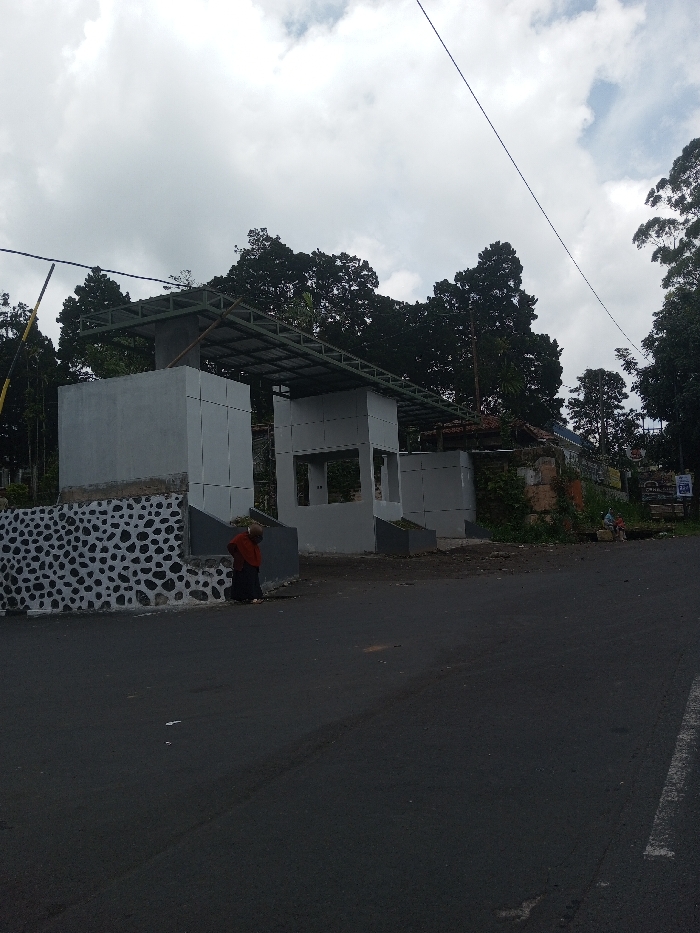The image depicts a roadside scene in Ura-Dozhi, Bhutan, under an overcast sky. In the foreground, a dark asphalt road, slightly marked with tire impressions, occupies the lower portion of the frame. To the left, a multi-part structure appears to be under construction or renovation, featuring light gray concrete walls and an unfinished metal framework roof. The base of the leftmost section of this structure is adorned with a decorative pattern of dark gray and white stones.
A single individual, dressed in a dark garment with a reddish-brown upper covering, stands near the base of the leftmost structure, facing away from the camera. Further to the right, behind the central portion of the structure, an older building with a tiled roof is partially visible. Adjacent to this, in the far right background, several indistinct figures are seated or standing near what appears to be a small roadside vendor or stall, identifiable by signs and some objects on the ground.
The background is dominated by dense, dark green trees and foliage, suggesting a heavily vegetated area. Overhead, utility lines traverse the sky diagonally from left to right. The overall impression is of a semi-urban or rural roadside area undergoing development.