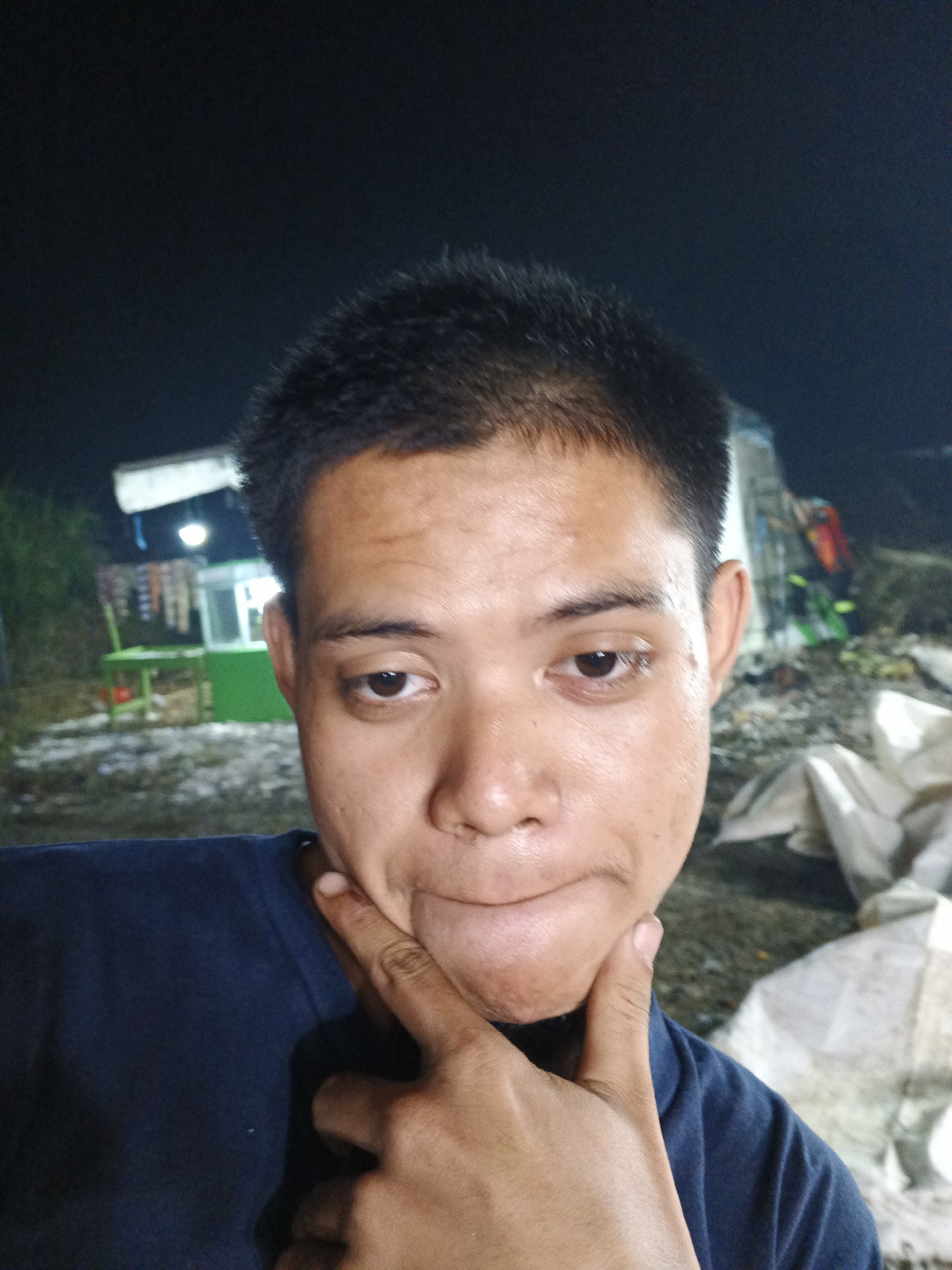 A close-up selfie of a young man with short dark hair and a dark blue t-shirt, taken at night. He is resting his chin on his hand, with his index finger on his cheek and thumb on his jawline. His eyes are open and looking towards the camera. In the background, a brightly lit food stall with green and white accents is visible, along with some hanging items. The ground appears to be dirt or gravel, and there are large white bags or bundles in the far right background. The overall setting suggests an outdoor, possibly market or roadside location, in Sukabumi, Indonesia.