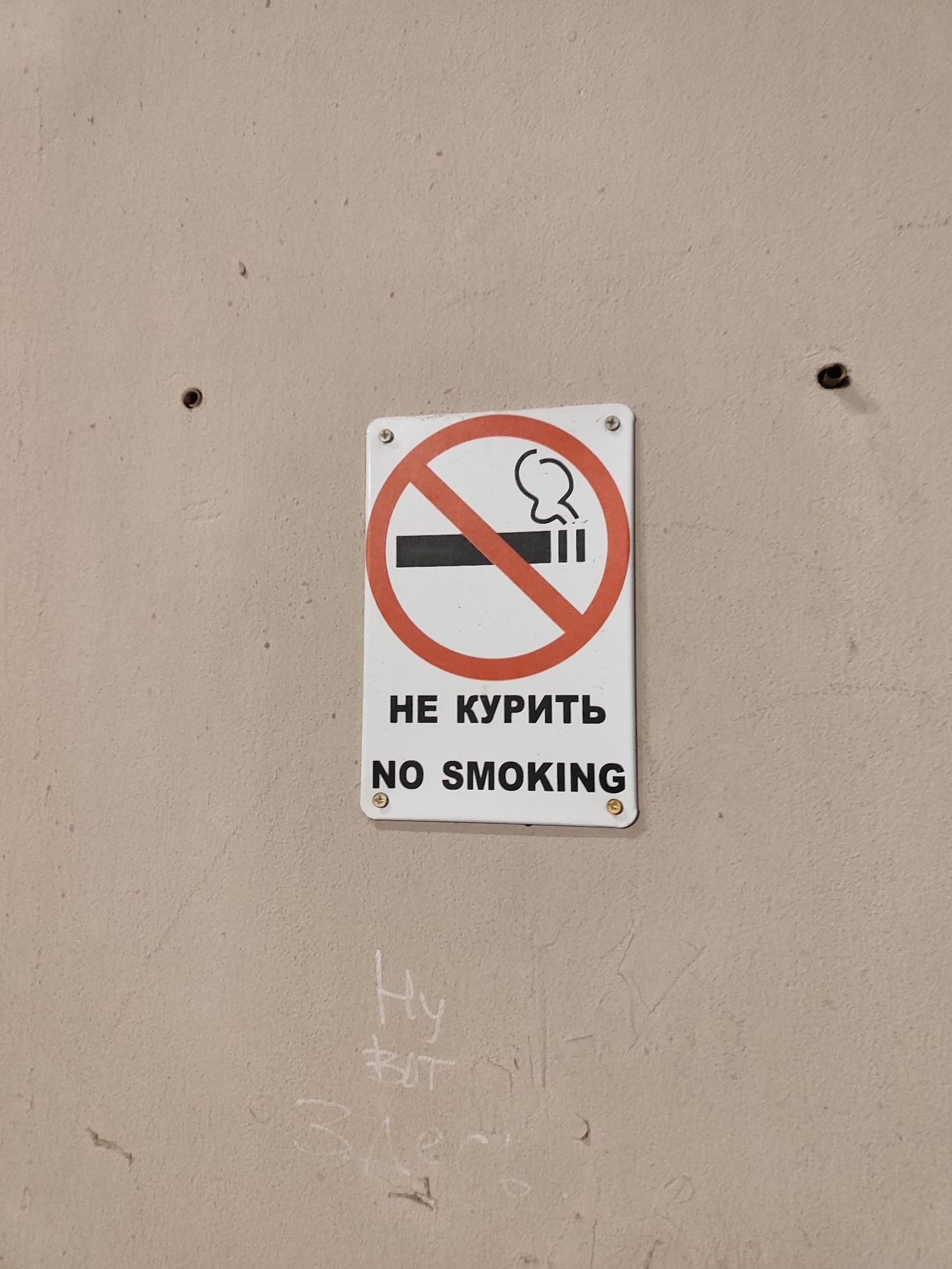 A white rectangular "No Smoking" sign is mounted on a textured, light beige or off-white wall. The sign features a standard international "no smoking" symbol: a black cigarette with a small smoke plume, enclosed within a red circle intersected by a diagonal red line. Below this graphic, the sign displays the warning in two languages: "НЕ КУРИТЬ" (Russian for "NO SMOKING") and "NO SMOKING," both in black capital letters. The sign is secured to the wall with four screws, one in each corner.
Above and to the left of the sign, a dark, small hole is visible in the wall, and another similar hole is present above and to the right, suggesting previous fixtures. The wall itself appears to have a slightly rough, granular texture and shows some minor marks and imperfections. Below the sign, faint, light-colored graffiti or etchings are visible on the wall. These include what appear to be the Cyrillic letters "Ну," some indistinct marks, the word "ROT" or Cyrillic "Рот," and the number "3." The lighting is even, indicating an indoor setting. No specific activity is depicted, only the static sign and wall.