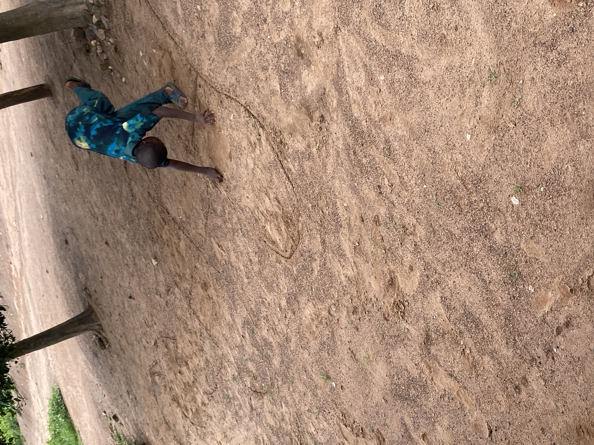 In an outdoor, sandy environment, a young boy wearing a blue and green tie-dye outfit and dark sandals is performing a gymnastic-like move. His body is angled towards the ground, with his hands on the dirt and his legs extended in the air. The ground is a mix of sand and small stones, with sparse green vegetation growing in patches. There are two trees visible in the background, casting shadows on the ground. The overall impression is of a child practicing or playing in a rustic, natural setting.