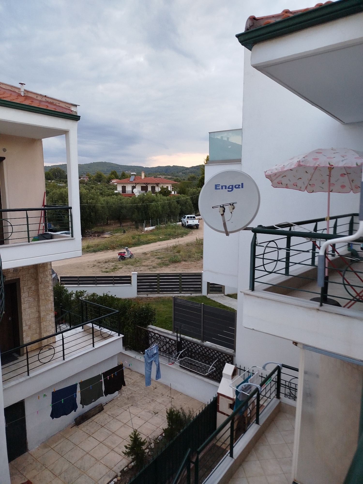 The image depicts a view from a balcony in Sithonia Municipal Unit, Greece. In the foreground, a white building with balconies is visible. On one balcony, a satellite dish with the brand "Engel" is mounted, alongside a pink and white patterned umbrella. Laundry is hanging on a clothesline in the courtyard below, including a pair of blue jeans and three dark-colored garments. In the middle ground, a dirt path winds through a grassy area with scattered trees. A red scooter is parked on the path, and a white pickup truck is driving away. In the background, a large white house with red-tiled roofs is nestled among olive trees, with green hills rising behind it under a cloudy sky.
