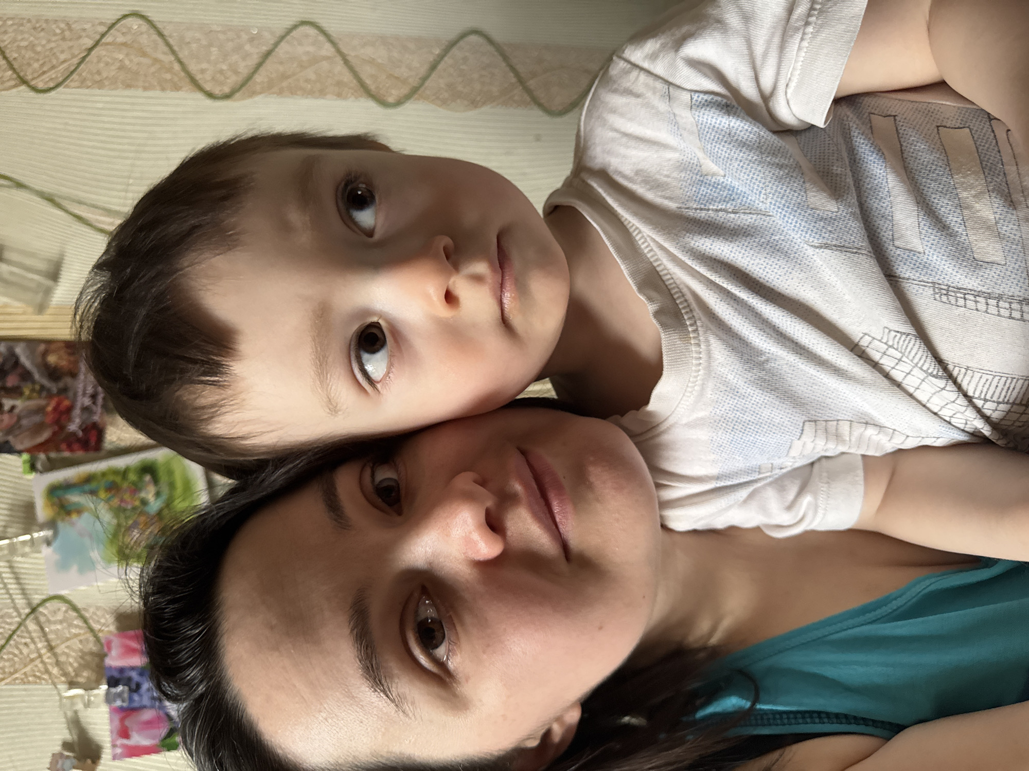 A close-up, intimate portrait features an adult woman and a young boy, likely a mother and child, captured in an indoor setting. The image is rotated 90 degrees clockwise, showing the subjects on their sides. The young boy's head is gently resting against the woman's forehead or temple, indicating a close bond. Both individuals are looking directly at the camera with calm, soft expressions.
The boy has light brown hair and wide, dark brown eyes. He is wearing a light-colored t-shirt, possibly white or off-white, which features faint, light blue and grey patterns that appear somewhat geometric or like stylized, indistinct lettering. The woman has dark hair and dark eyes, and she is wearing a teal or dark turquoise-colored top.
The background is softly lit and consists primarily of a light-colored patterned textile, possibly bedding or a blanket, with a repeating wavy or zig-zag motif in a muted green hue. In the upper left background, behind the woman's head, some blurry, colorful rectangular shapes are visible, which could be small pictures or decorative items affixed to a wall or surface. The lighting is soft and even, suggesting a daytime indoor setting. There is no discernible text visible in the image.