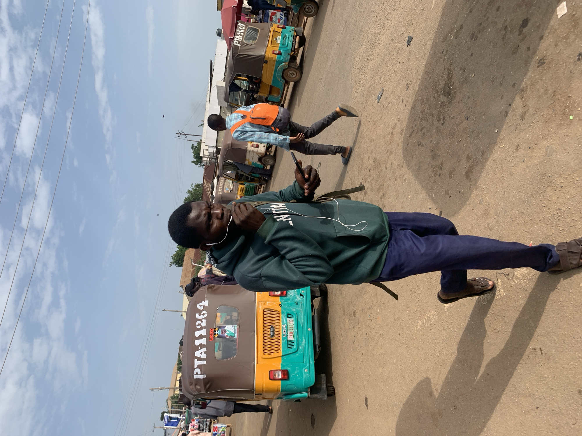 In Gura Top, Nigeria, a street scene unfolds with several auto-rickshaws parked and people moving about. In the foreground, a young man walks toward the right, wearing a dark green hooded sweatshirt, blue pants, and brown sandals. He holds a smartphone in his right hand and appears to be speaking into earbuds. Behind him, an auto-rickshaw with "PTA 11264" displayed on its roof is visible. Further in the background, another young man wearing a light blue checkered shirt and an orange backpack walks away from the camera. Other auto-rickshaws and a few more individuals are scattered throughout the scene, suggesting a bustling urban environment. The ground is unpaved, and overhead power lines are present, indicating the presence of infrastructure. The sky is a pale blue with scattered clouds.