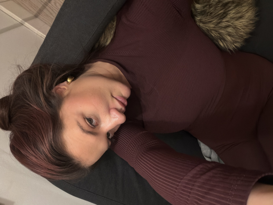 The image captures a close-up of a woman lying down, with her head resting on a dark surface, likely a couch or chair. The image is oriented such that the woman's head is on the left side of the frame, and her body extends towards the right.
The woman has dark brown hair with visible reddish or auburn highlights, pulled back into a bun at the back of her head, though some wisps frame her face. Her complexion is smooth, with a natural makeup application that includes a touch of blush on her cheeks. Her eyes are open, dark, and she is looking directly at the camera. Her lips are slightly parted, revealing a natural, almost neutral expression with a hint of a soft smile. A small, golden, possibly dome-shaped or hoop earring is visible in her left ear.
She is wearing a form-fitting, long-sleeved garment in a dark burgundy or maroon color. The fabric has a distinctive ribbed texture, visible throughout the sleeves and torso. The garment appears to be either a top or a jumpsuit, hugging her form.
Behind her head and upper body, part of a fluffy, light brown or beige object, possibly a faux fur pillow or throw, is visible. The immediate surface she is resting on is a dark gray or black fabric, consistent with upholstery. The overall lighting suggests an indoor setting, possibly with soft, warm artificial light. There is no visible text in the image.