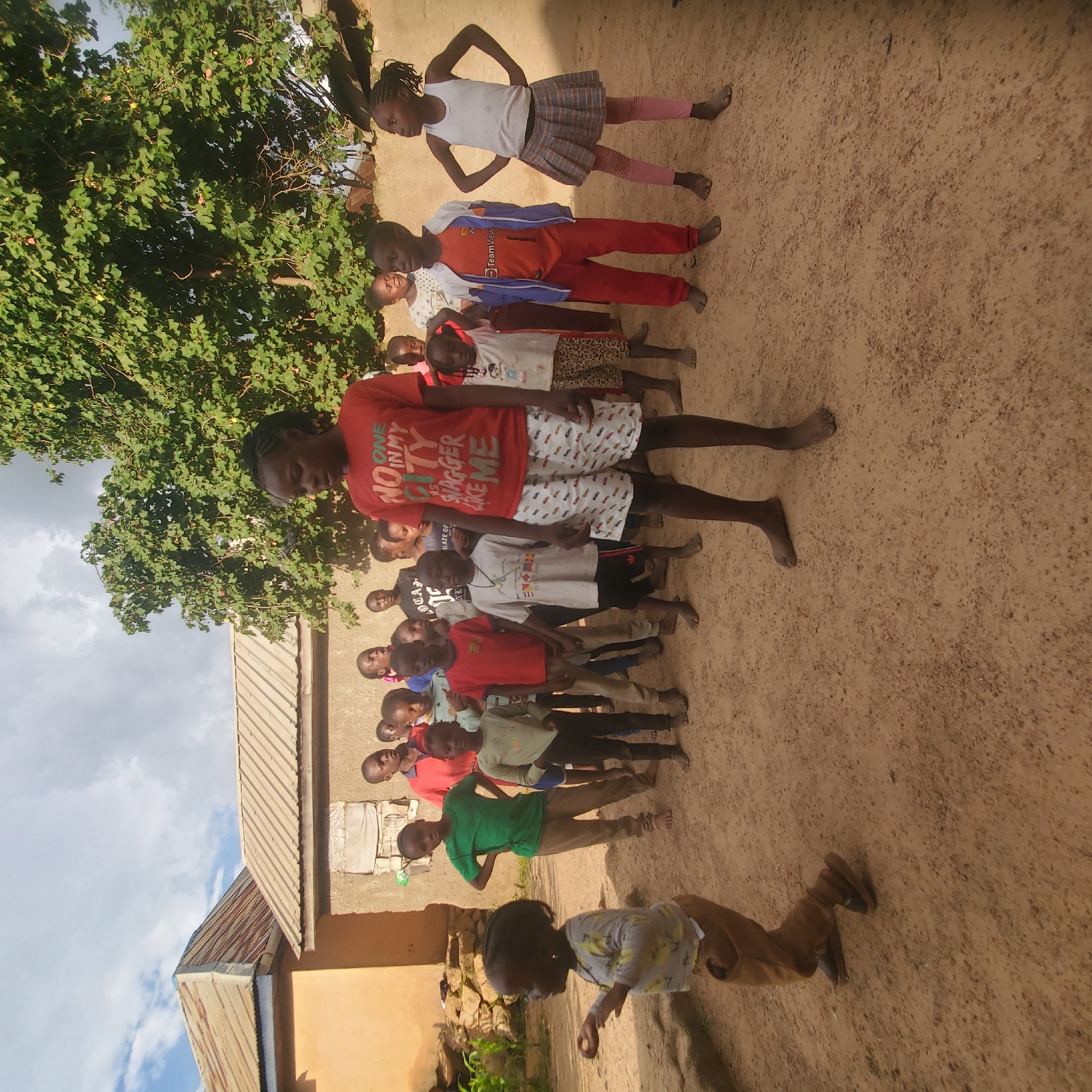 In an outdoor scene in Jos, Nigeria, a group of children stands on a dusty, sandy ground. A large green tree with lush leaves provides shade in the background, alongside two simple buildings with corrugated metal roofs. In the foreground, a young girl in a light-colored t-shirt with a yellow pattern and brown pants is captured mid-motion, her arms bent as if about to throw or catch something. Several other children are visible, some in the foreground and others further back, all appearing to be participating in an activity or waiting for instruction. They are dressed in casual, everyday clothing. The sky above is partly cloudy with patches of blue.