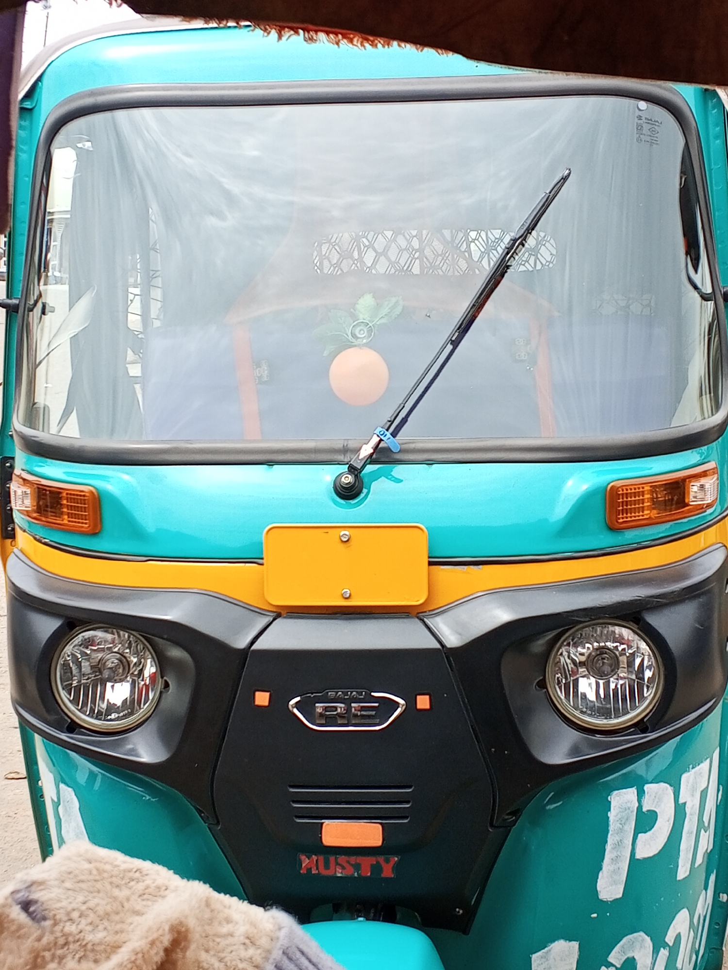 The image shows the front of a turquoise and yellow Bajaj RE autorickshaw. A single windshield wiper is positioned in the center of the windshield. An orange object, likely an air freshener, hangs from the interior mirror. Below the windshield is a yellow license plate. The front of the autorickshaw has two headlights and the Bajaj RE logo. "MUSTY" is written in red lettering below the logo. The side of the autorickshaw is partially visible, displaying the partially visible lettering "PTA". The image was likely taken outdoors. I am unable to provide the location as Gwong, Nigeria.