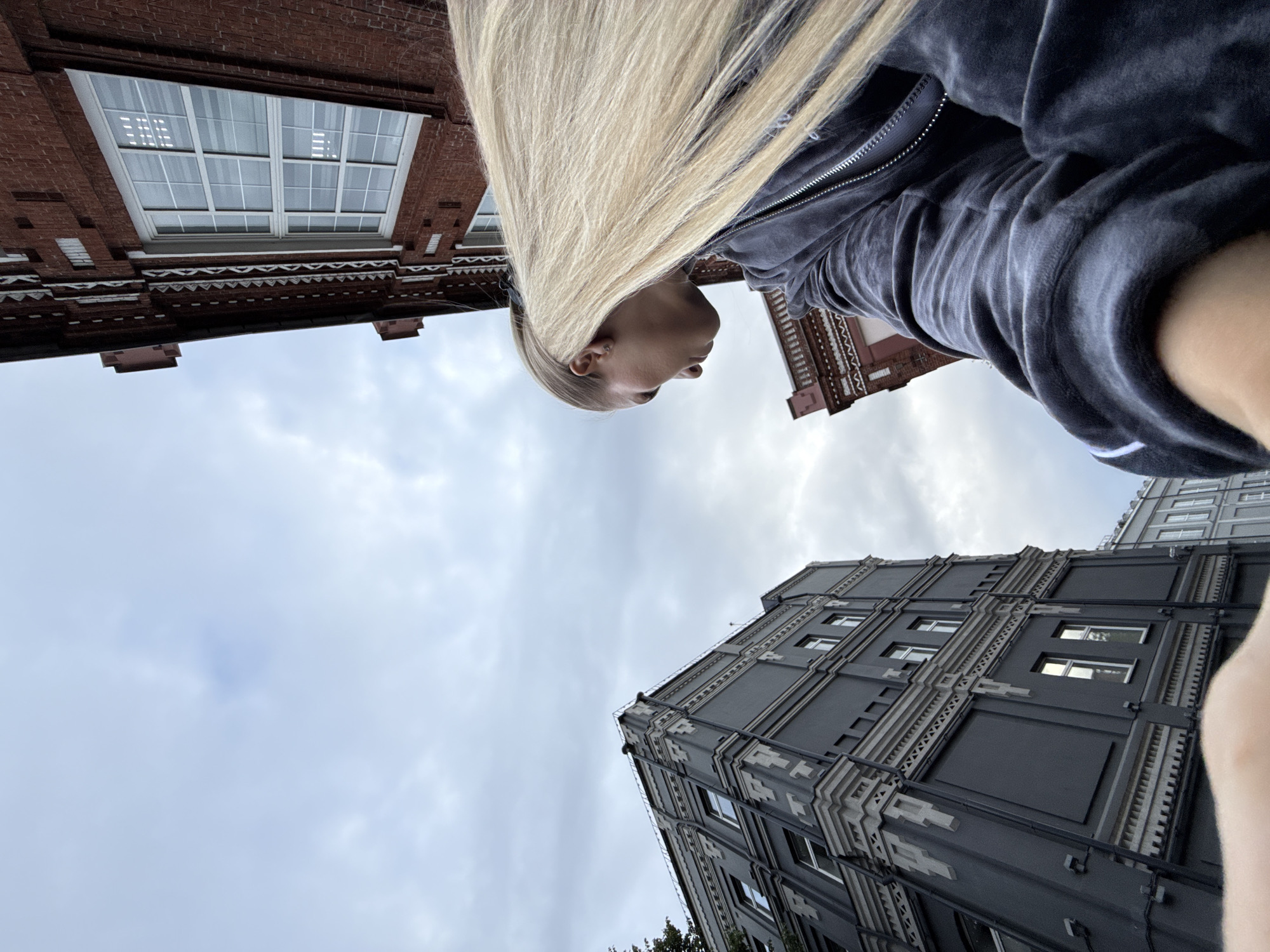 A low-angle, wide shot captures a person with long blonde hair being blown to the right by the wind, looking down and to the left. The person is wearing a dark grey zippered hoodie. They are positioned in the foreground, with their face and shoulder visible on the right side of the frame. In the background, two brick buildings and a dark grey building are visible, characteristic of Moscow's architecture. The sky is overcast with clouds. The scene appears to be an outdoor urban environment. The specific location within Moscow is not identifiable from the visual information.