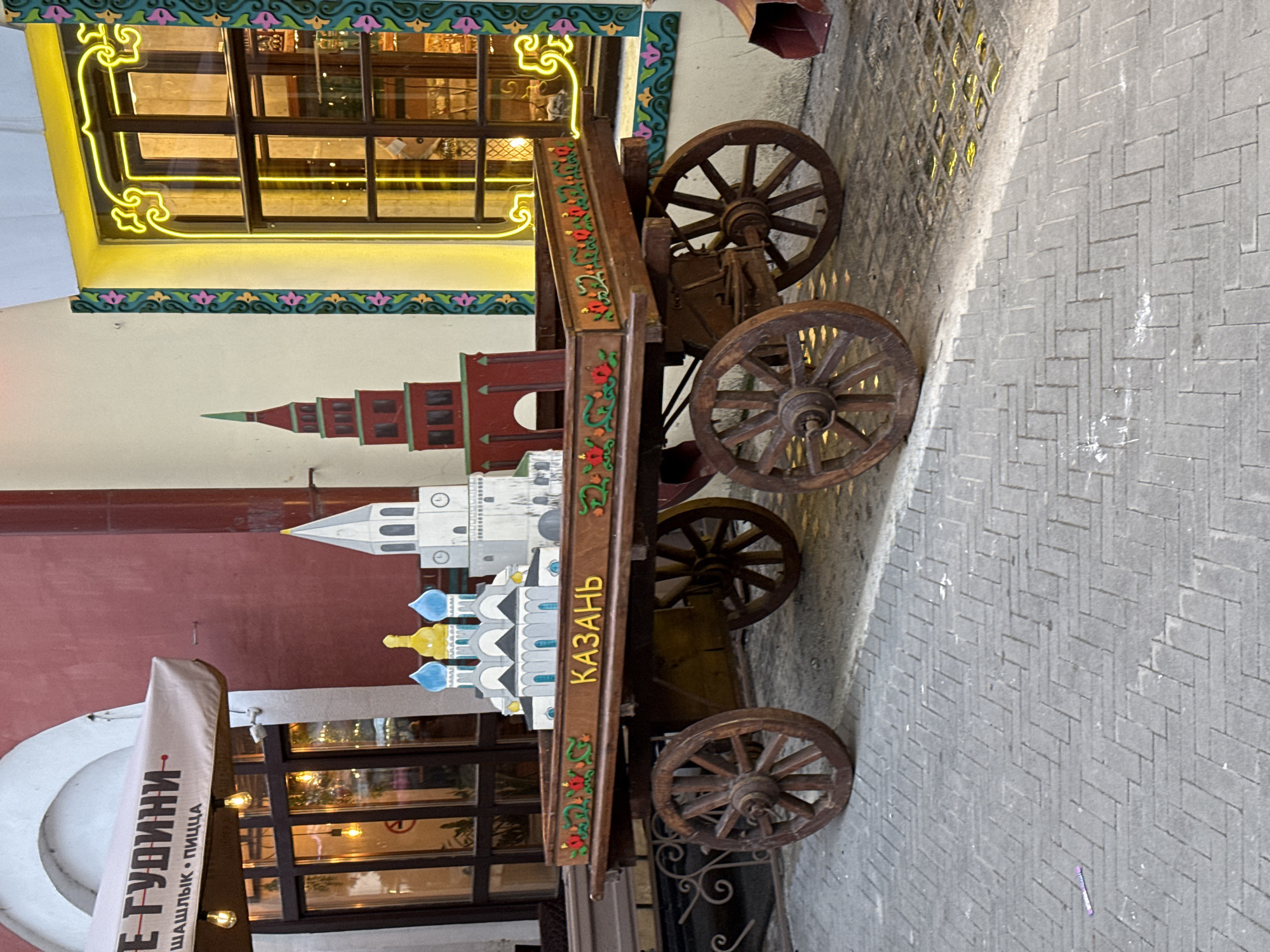 An outdoor scene features a decorative wooden cart positioned on a paved ground, likely a street or alleyway in Kazan, Russia. The cart has four large, spoked wooden wheels and its long, rectangular side is adorned with vibrant red and green floral patterns. The word "КАЗАНЬ" (KAZAN) is prominently displayed in gold lettering on the cart's side.
Perched atop the cart are miniature architectural models, depicting significant landmarks of Kazan. These include a red-colored tower resembling the Söyembikä Tower, a white building with a tall spire, and another building featuring distinctive blue and yellow domed roofs, likely representing the Qolşärif Mosque or a cathedral.
In the background, building facades are visible. One building exhibits a colorful decorative trim with green, pink, and purple patterns, and a window framed by a glowing yellow neon outline. Another building has a reddish-brown wall.
To the left, a partially visible white banner or flag hangs from an awning. Visible text on the banner reads "Е ГУДИНИ" (E GUDINI) and below it, "ШАШЛЫК • ПИЦЦА" (SHASHLIK • PIZZA), indicating a restaurant or cafe. The ground is paved with grey rectangular bricks arranged in a herringbone pattern, with some light reflections visible. No people are present in the image. The lighting suggests it could be daytime, though the neon and reflections hint at artificial light or an evening setting.