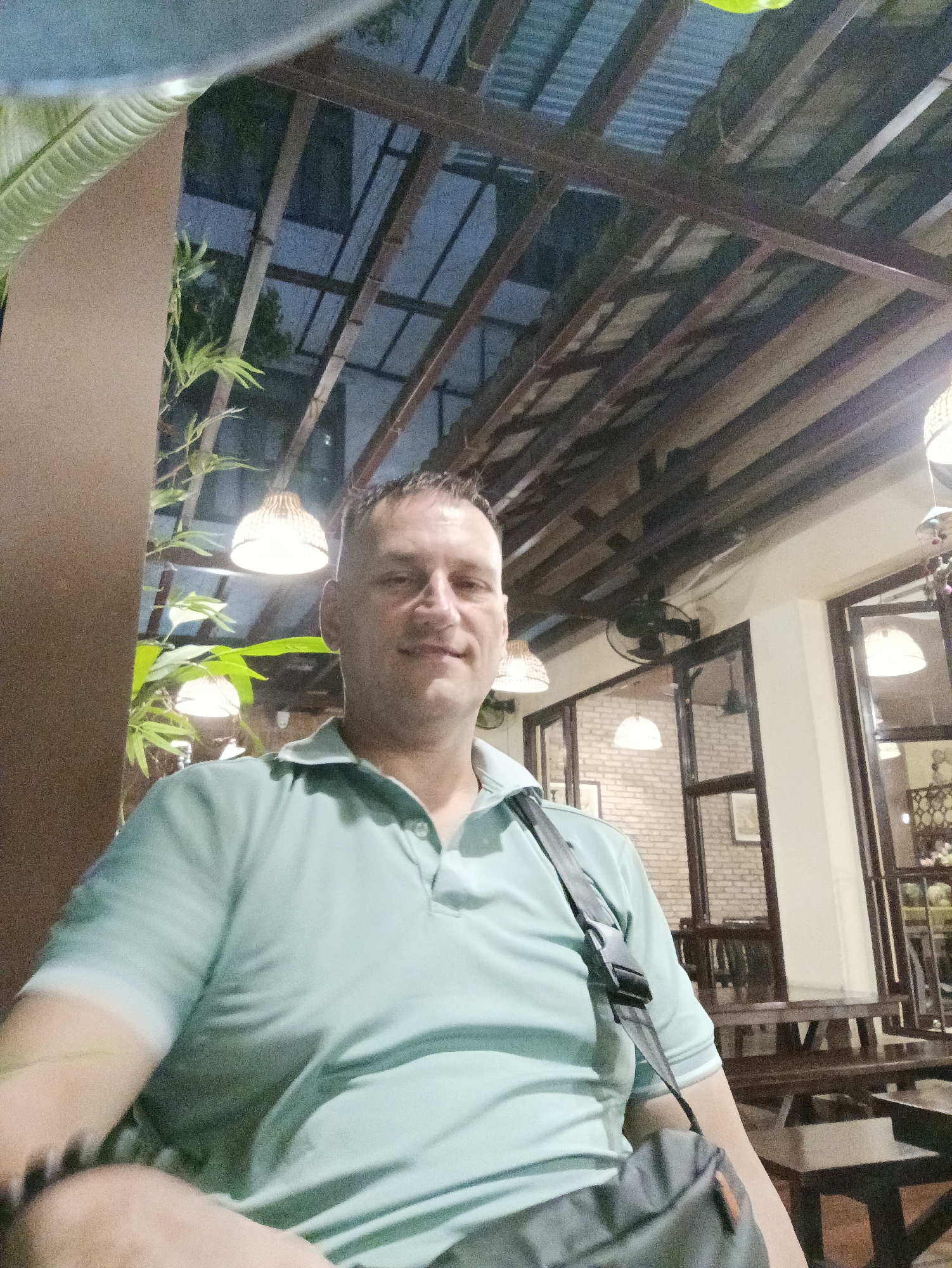 The photograph captures a man in a light green polo shirt, seated in an outdoor or semi-outdoor setting. He wears a dark-colored bag slung over his shoulder. The structure above is made of metal supports. Lamps are hanging down on the ceiling. The location is Nha Trang Ward, Vietnam. The scene includes tables and chairs. The environment suggests a restaurant or cafe setting. The time appears to be nighttime or early evening, with artificial lighting illuminating the scene. Behind the man, a wall features windows and a brick facade.