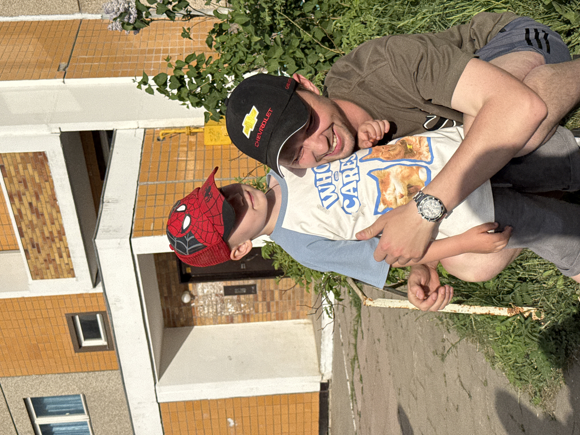 A man and a child are outdoors near a multi-story building in Chaikovsky, Russia. The building has yellow brick and white trim. The man wears a black baseball cap with a Chevrolet logo, a brown t-shirt, grey shorts, and a watch on his left wrist. He is lying down and hugging the child. The child wears a red and black Spiderman cap and a white t-shirt with blue sleeves. They are on a patch of grass near a sidewalk.