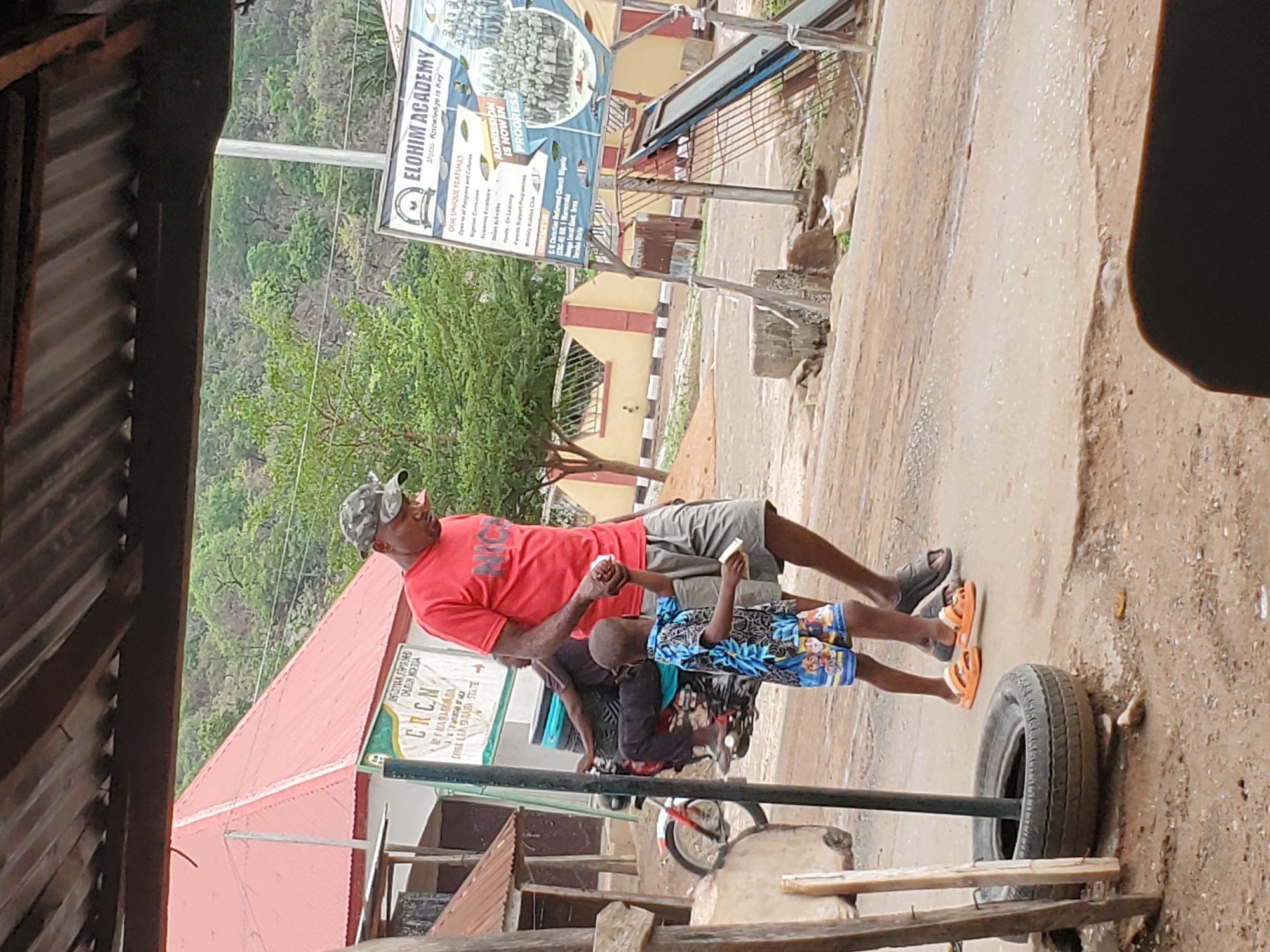 The image shows a man in a red shirt and camouflage hat holding hands with a child wearing a blue patterned shirt and shorts. They stand on a dirt path alongside a tire, with a wooden structure in the foreground. In the background, signs indicate "ELOHIM ACADEMY" and "CRCN MARARABA, DONEA LEA DARABA STATE," placing the location in Mararaba, Nigeria.