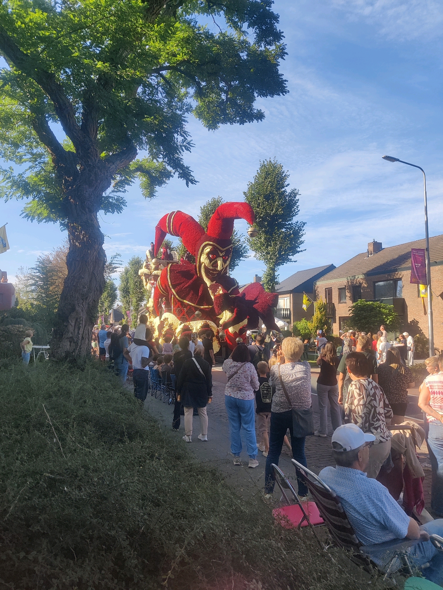 The image captures a parade float, resembling a jester, passing through a street lined with spectators in Zundert, Netherlands. The float is elaborately decorated with red and gold fabric, forming a large jester figure with a distinctive hat. A crowd of people of various ages stands on the sidewalk, watching the procession. Some individuals are seated in folding chairs, while others stand or are being carried on shoulders. The street is paved, and houses are visible in the background, some adorned with banners. Lush green trees line the street, casting shadows on the scene. The sky is a clear blue with wispy clouds, and a streetlamp is present on the right side of the image. The overall atmosphere appears festive, indicative of a community event or celebration.