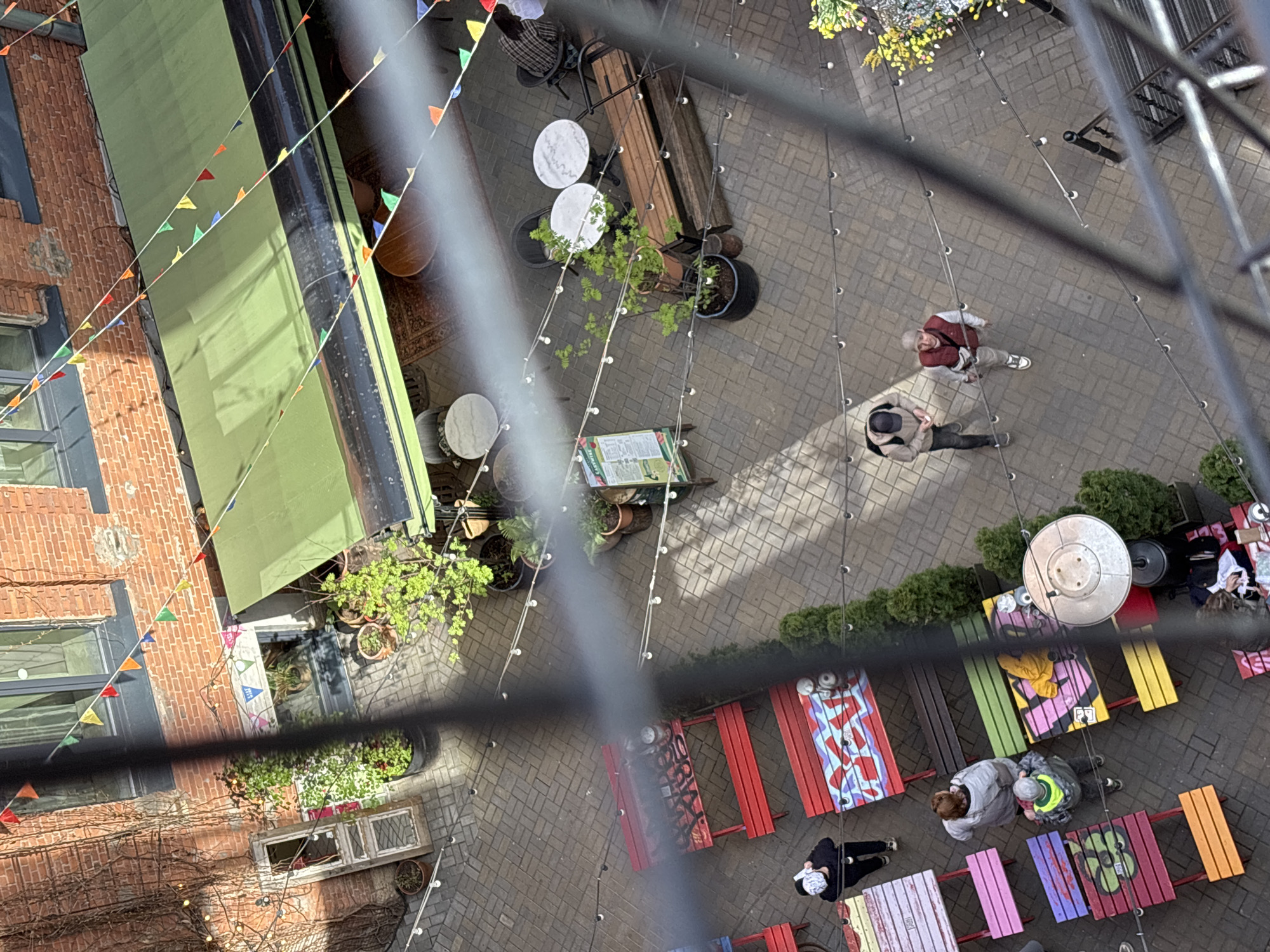The image depicts an outdoor courtyard in Saint Petersburg, Russia, viewed from above, likely from a high window or balcony. The courtyard is paved with light-colored bricks and contains several seating areas with colorful picnic tables and chairs. Several people are present, some walking and some seated at the tables. A green awning extends over one side of the courtyard, and colorful triangular flags are strung across the space. Potted plants and greenery are scattered throughout the area, adding to the ambiance. The architecture surrounding the courtyard is characterized by brick buildings with windows. A metal structure, possibly a part of a balcony or railing, is visible in the foreground, partially obscuring the view. String lights are strung throughout the courtyard.