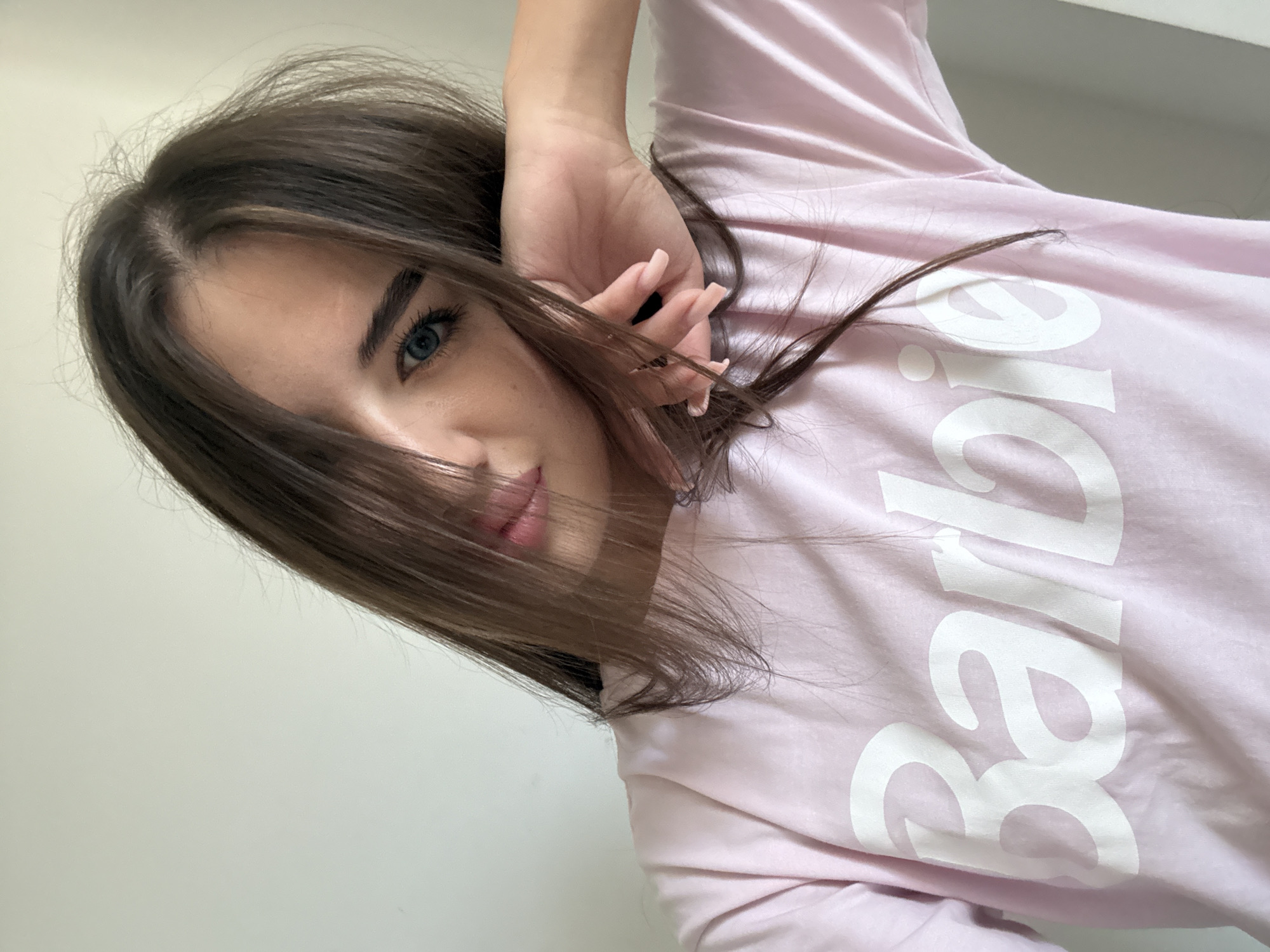 A young woman with long, dark brown hair and blue eyes is captured in a close-up, upper-body selfie. She is wearing a light pink t-shirt with the word "Barbie" printed in white, stylized letters across the chest. Her hair is partially obscuring her face, with strands falling across her forehead and cheek. She is resting her right hand against her face, with her fingers gently touching her cheek. The background is a plain, light-colored wall, suggesting an indoor setting. There are no discernible features that indicate a specific location in Chișinău, Moldova. The lighting is soft, likely from an overhead source or natural light.