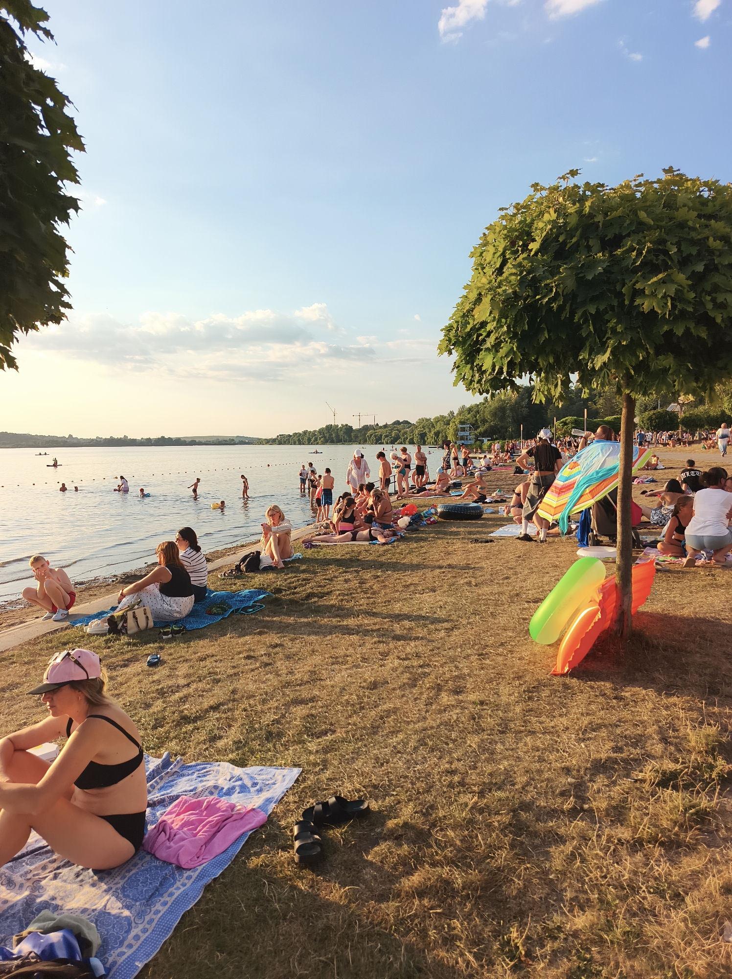The image depicts a sunny day at a beach, likely located in Ternopil, Ukraine, given the context. A body of water, possibly a lake or river, is visible in the background, with people swimming and wading. The shore is lined with a grassy area where numerous individuals are relaxing on towels and blankets. Some people are sunbathing, while others are engaged in conversation or looking at their phones. In the foreground, a woman in a black bikini and a pink cap is seated on a blue and white patterned towel, with a pink towel and a black pair of sandals beside her. Further to the right, a tree with green foliage stands, and near its base are two inflatable pool toys, one green and one orange. In the mid-ground, an umbrella with colorful stripes is visible. The sky is blue with scattered white clouds, and the sun appears to be setting or rising, casting a warm glow on the scene. There are trees and buildings visible on the far bank of the water.