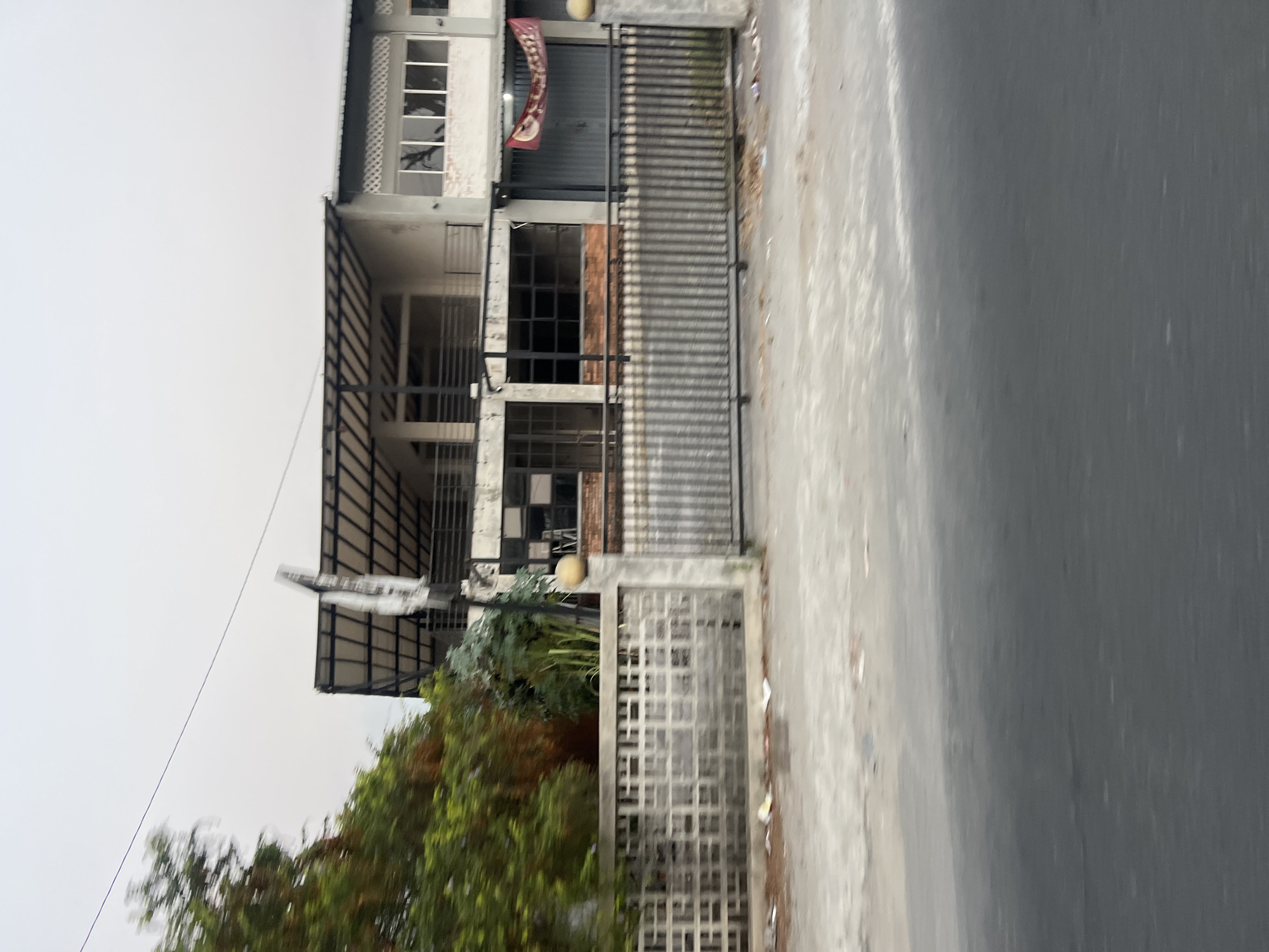 This is a medium-shot, eye-level, horizontal, slightly blurred outdoor photograph of a multi-story building with a large open balcony on the second floor and a metal canopy extending over the ground floor. The building appears to be under construction or renovation, with visible brickwork on the ground floor and unfinished exterior walls. A dark metal fence with vertical bars stands in front of the building, separating it from a paved area and a road. A portion of a patterned concrete wall is visible to the left of the metal fence. Lush green trees and foliage are situated to the left of the building and fence, with a utility pole and wires visible in the upper portion of the frame. The sky is overcast and appears to be a pale gray. A red banner is partially visible hanging on the right side of the building. The scene is located in Sendangadi, Indonesia.