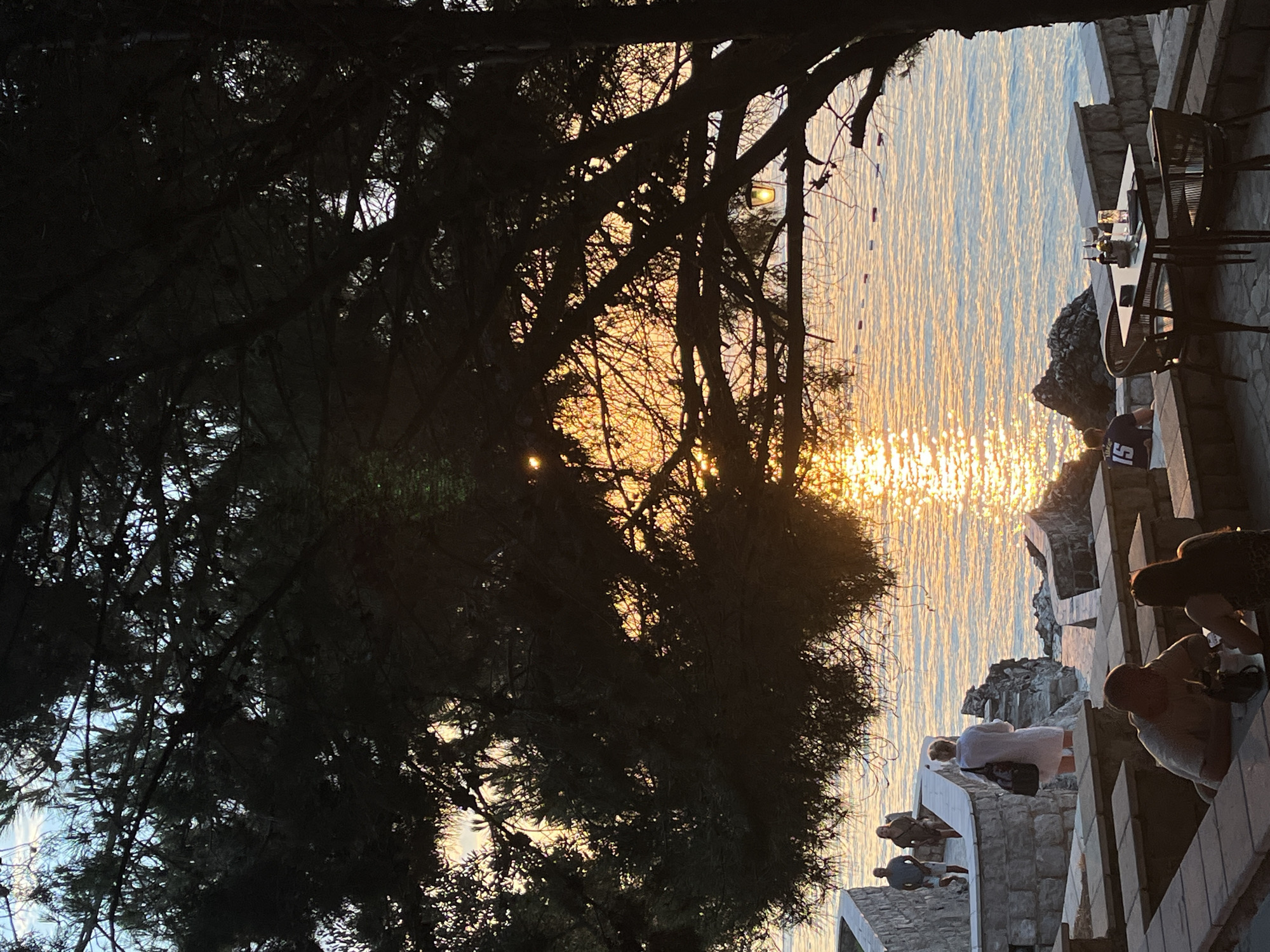 In the city of Pečurice, Montenegro, a sunset scene unfolds by the Adriatic Sea, viewed from a stone terrace shaded by a large pine tree. The setting sun casts a golden, shimmering reflection across the tranquil water, with a few buoys marking a swimming area in the distance. On the terrace, a table is set with two chairs, water glasses, and condiment bottles, awaiting patrons. Further back, several people are visible. Two men and a woman walk along a stone pathway leading to the water's edge, while another woman in a white tunic stands near a stone staircase. In the foreground, a couple sits at a stone bench, seemingly engaged in conversation. The overall atmosphere is serene and inviting, capturing a peaceful evening by the sea.