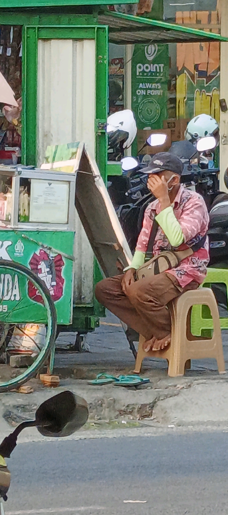 A man wearing a pink camouflage-patterned shirt, light green arm sleeves, brown pants, a black baseball cap, and a face mask sits on a beige plastic stool on a sidewalk in Sidoarum, Indonesia. The man is covering his face with his right hand, and he has a beige messenger bag slung across his chest. Next to him is a green food cart with a bicycle wheel visible on its left side. Behind the man, there are several motorcycles with helmets. The ground is paved with concrete, and a pair of turquoise flip-flops are on the ground in front of the stool. A motorcycle side mirror is visible in the foreground, on the left side of the image. The scene appears to be a street in a commercial area, possibly a market.