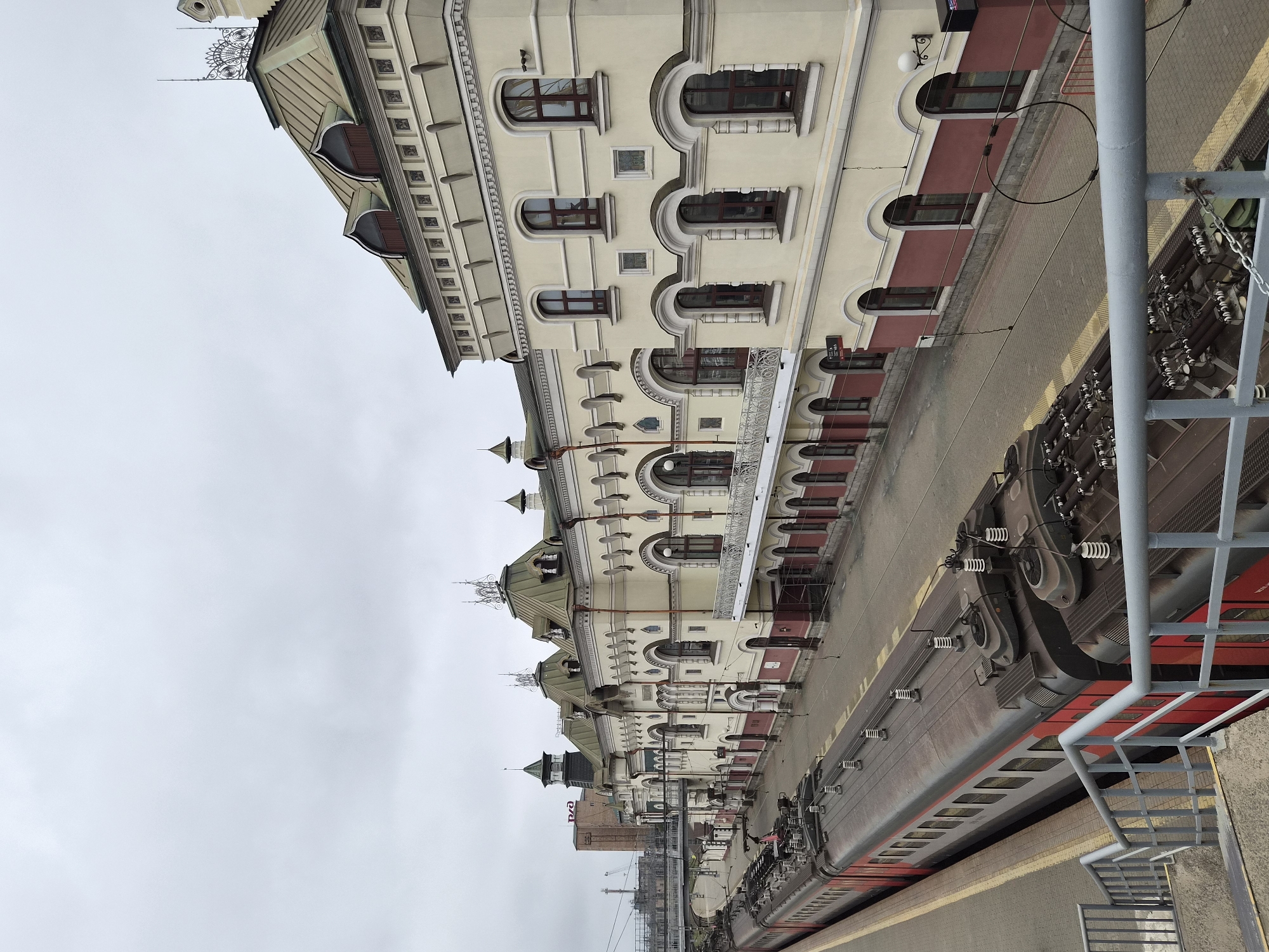 The image shows a view of the Vladivostok railway station on an overcast day. The grand, historic station building is painted in cream and burgundy, with ornate architectural details, multi-paned windows, and a green-tiled roof topped with decorative spires. A red and grey passenger train is parked at the platform alongside the building. The platform is paved with yellow and grey tiles, and a grey metal staircase with a railing leads down towards the tracks. In the background, industrial structures and a bridge are visible. The scene is devoid of people, focusing entirely on the architecture and the train.
