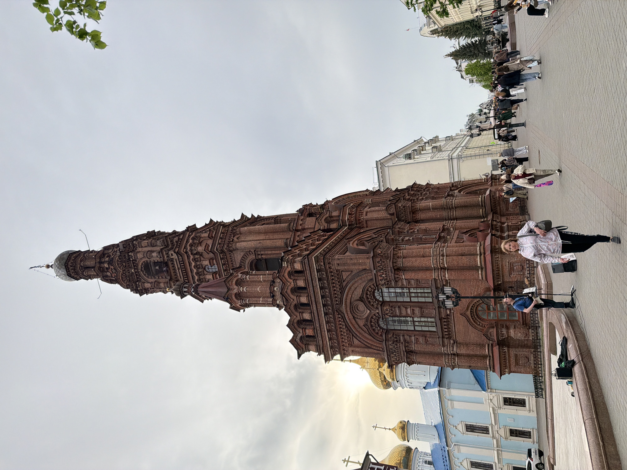 The image shows the Leaning Tower of Soyembika in Kazan, Russia, viewed from a low angle. The tower, made of red brick, stands prominently against a cloudy sky. A crowd of people is present on the cobblestone street beside the tower. Also visible are parts of a blue and white building with golden domes, typical of Russian Orthodox churches. A lamppost is present near the base of the tower. A man in a blue shirt and black pants is setting up equipment, presumably for musical performance. An older woman stands next to him.