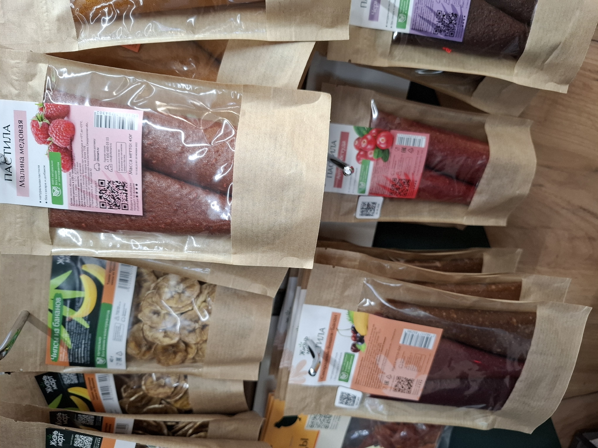 The image shows a close-up view of several retail packages of dried fruit products, likely a market or grocery store display. The packages are made of brown paper with clear plastic windows, revealing the contents. The products are labeled in Russian, with prominent text indicating "ПАСТИЛА" (pastila, a type of fruit leather) and various fruit flavors such as "Малина медовая" (honey raspberry), "Смородина-Вишня-Банан" (currant-cherry-banana), and "Черника" (blueberry). Some packages also show images of the fruits. The background is a light-colored wooden surface. The overall impression is a neat and organized display of food items. There is no discernible connection to the city of Tyumen, Russia, or any specific location within it. There are no people, actions, or interactions visible in the image.