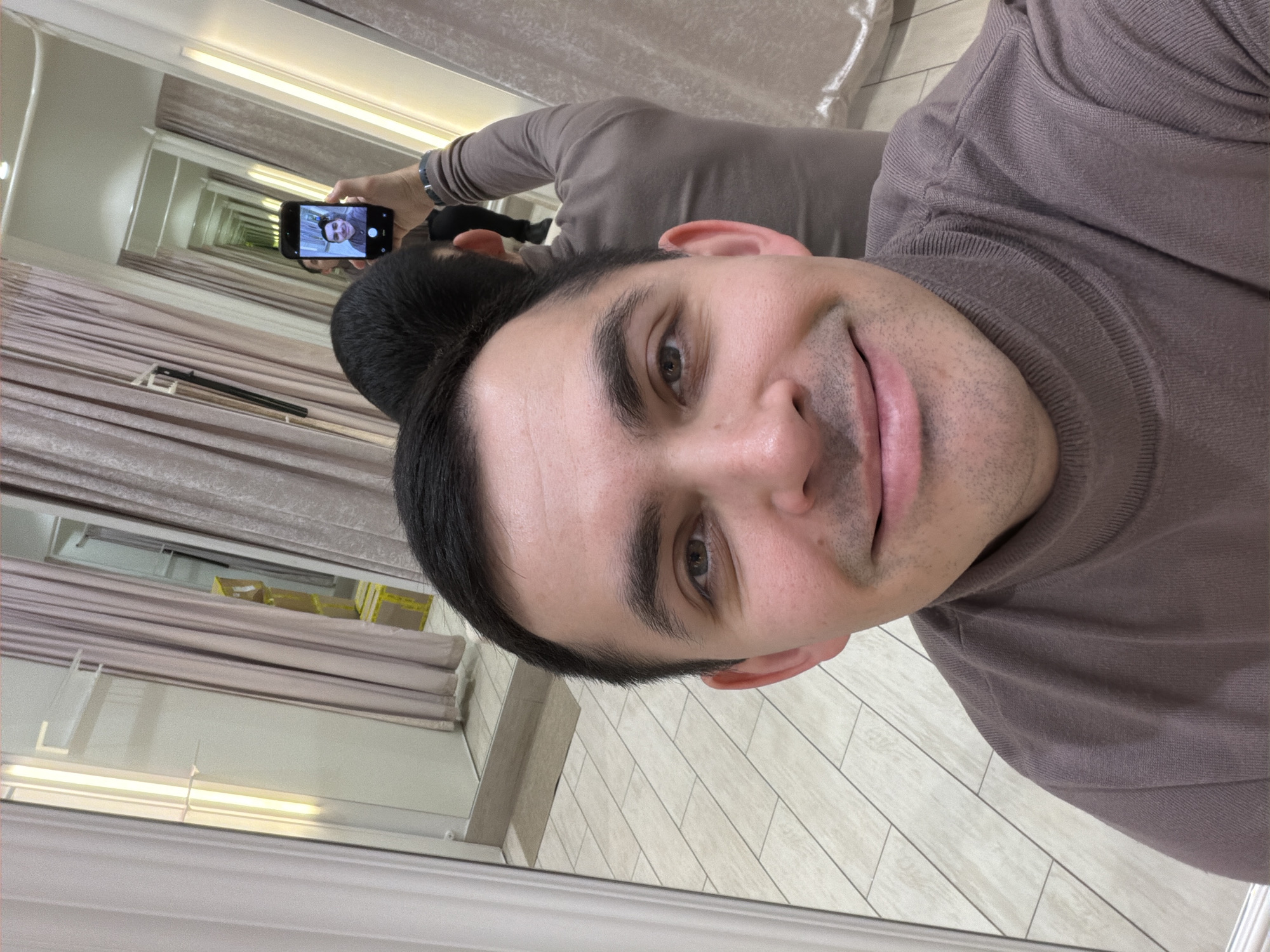 A man is taking a selfie in a mirror. He is wearing a brown turtleneck sweater. In the mirror, another image of him taking the selfie is visible, making it a picture-in-picture effect. The background features what appears to be a dressing room or retail space, with curtains and tiled flooring. The location is Izhevsk, Russia.