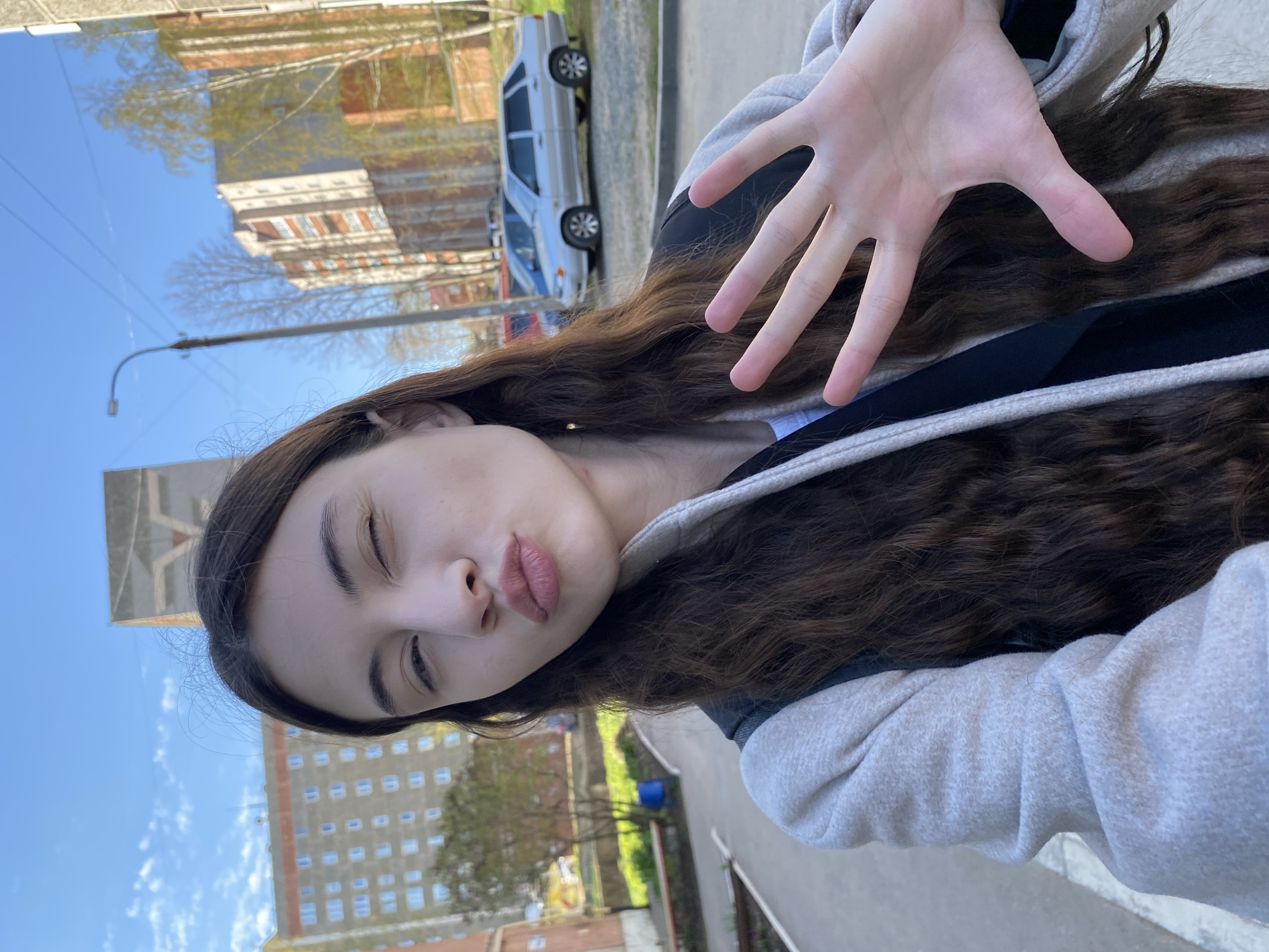 A young woman is taking a selfie outdoors in what appears to be a city setting, possibly Votkinsk, Russia given the building style in the background. She is partially facing the camera, with her head slightly tilted to the right. She has long, dark brown hair, and she is wearing a hoodie with a light grey exterior and a black interior. Her right hand is raised, with her fingers spread open toward the camera. In the background, there are several multi-story buildings, a car, trees, and a blue sky. The photo appears to be taken in the daytime.