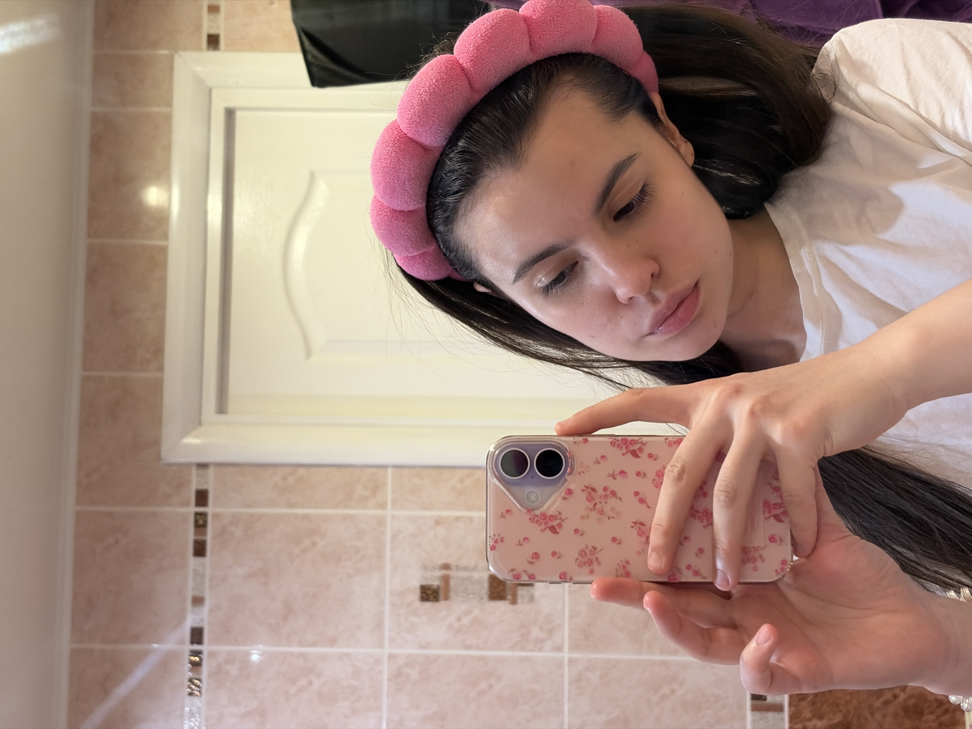 A woman with long dark hair, wearing a pink bubble headband and a white shirt, is taking a selfie in a bathroom. She is holding a pink phone with a floral case. The bathroom has light brown tiles with a decorative strip and a white door or cabinet. The city of Chaikovsky, Russia, is not discernible from the image.