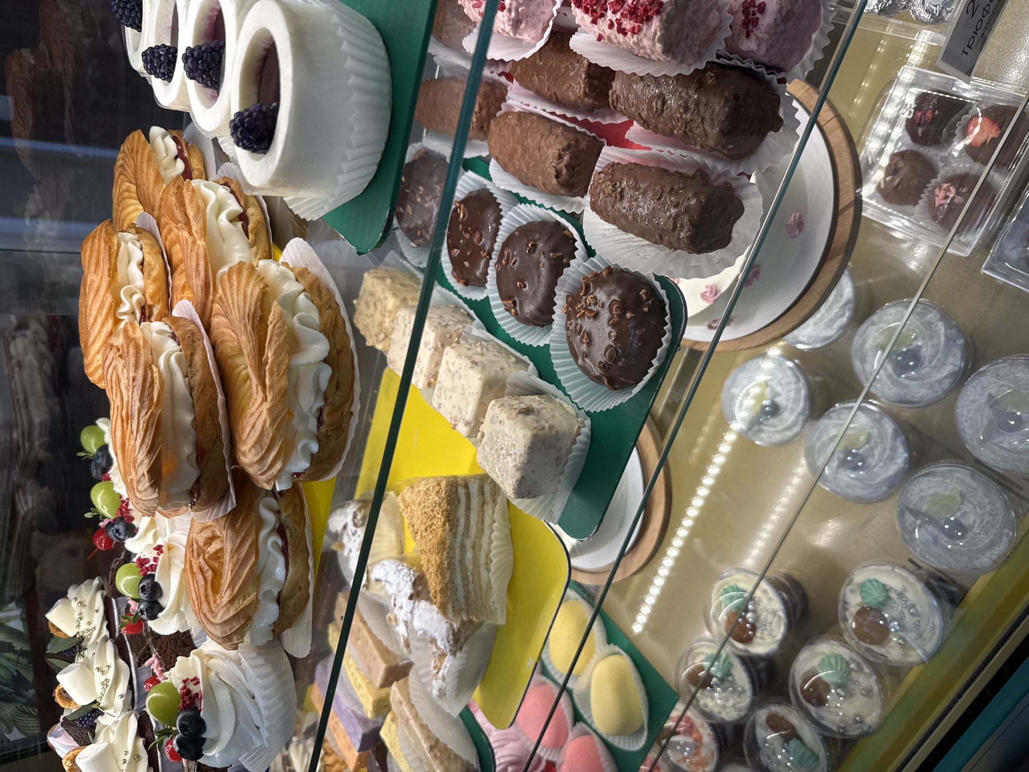 The image depicts a display case filled with various desserts, including cream-filled pastries, chocolate-covered sweets, and miniature cakes. The desserts are arranged on multiple tiers within the case. Based on additional sources, it is likely this is a scene from a bakery or confectionery in the city of Chaikovsky, Russia.