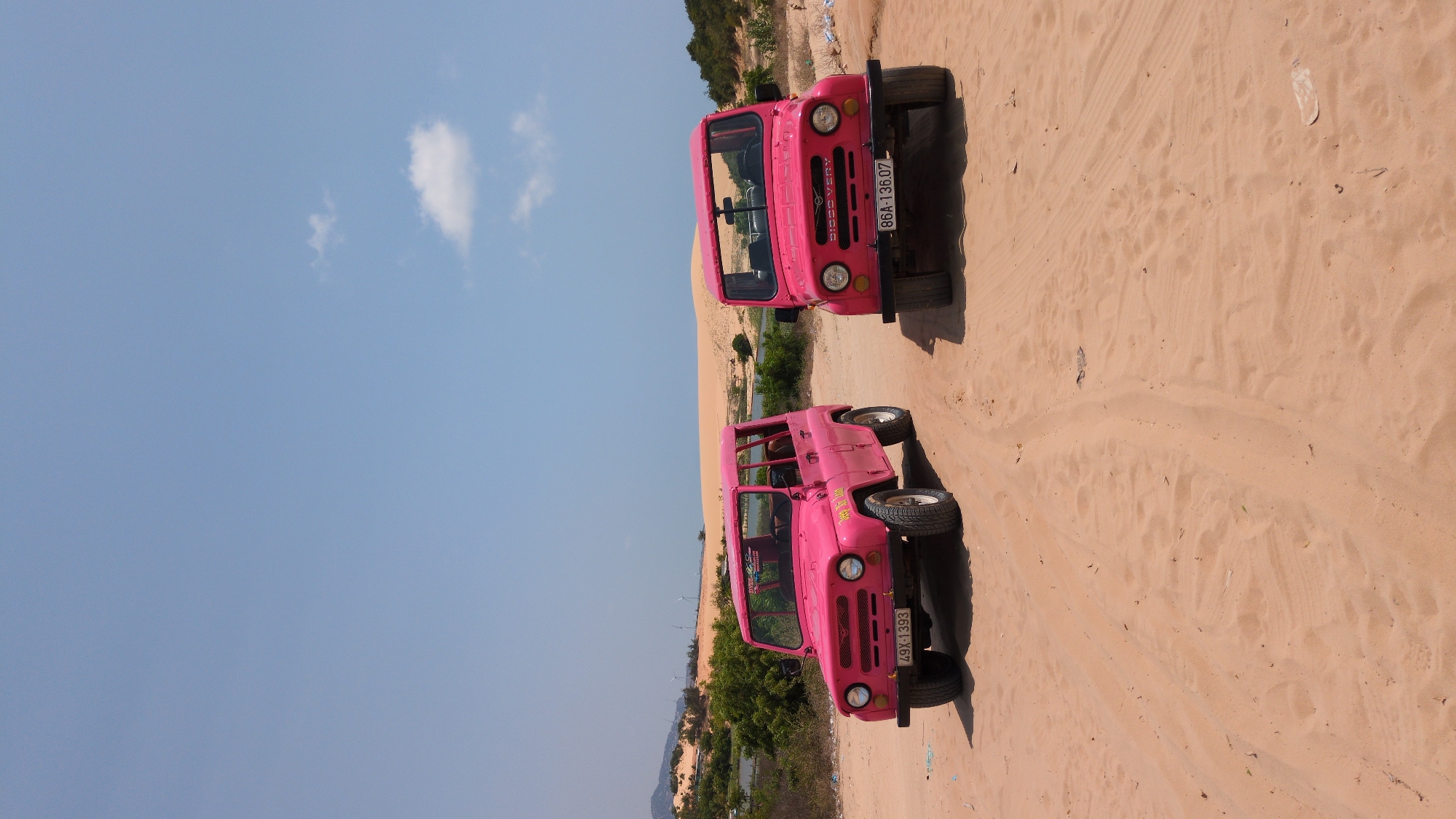The scene is a landscape in Phuoc Dinh, Vietnam, showcasing two pink off-road vehicles, likely jeeps, parked on a sandy terrain with visible tire tracks. The sky is clear and blue, with a few scattered clouds. Some sparse vegetation can be seen near the edge of the sand, suggesting a transition to a different ecosystem nearby. The vehicles appear to be oriented in slightly different directions relative to each other.