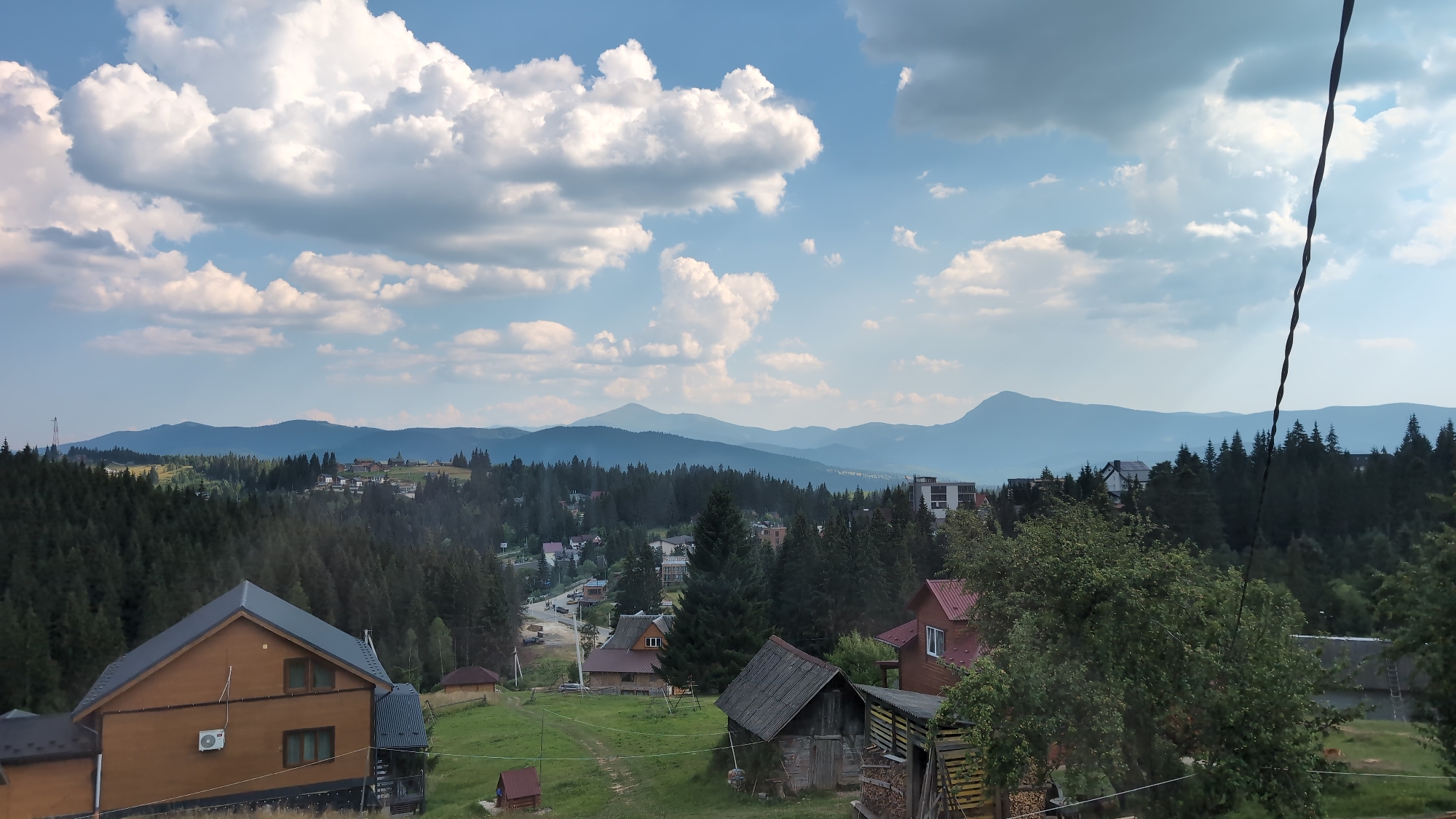The image depicts a panoramic view of a mountainous landscape in Yablunytsya, Ukraine, under a partly cloudy sky. In the foreground, several wooden houses with dark roofs are visible. One prominent brown wooden house features an outdoor air conditioning unit on its side. Adjacent to it is a smaller, rustic wooden shed. Further into the scene, more houses and buildings are scattered amidst dense coniferous forests that cover the rolling hills. A road winds through the village, with a few cars visible. In the distance, layers of blue mountains recede into the horizon, softened by atmospheric haze. The overall scene conveys a peaceful, rural atmosphere in a mountain resort area.
