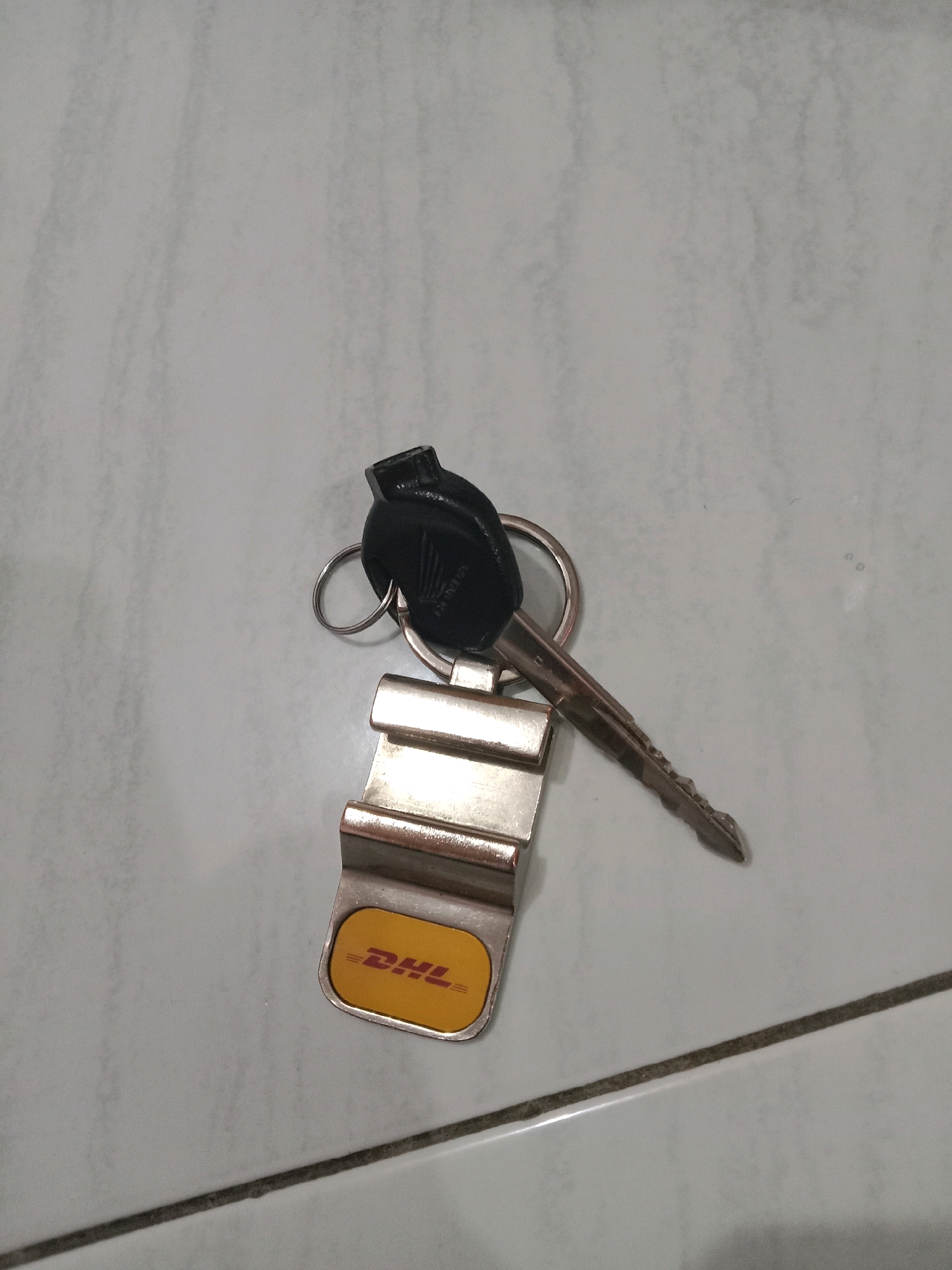A Honda key with a DHL keychain is lying on a tiled floor. The key is silver with a black plastic head. The DHL keychain is a rectangular metal tag with a yellow background and the red DHL logo. The floor is made of light-colored square tiles with dark grout lines. The lighting is dim, suggesting an indoor environment. The location is identified as Brangsong, Indonesia, though there are no visual cues in the image to confirm this specific location. No people are present in the image.