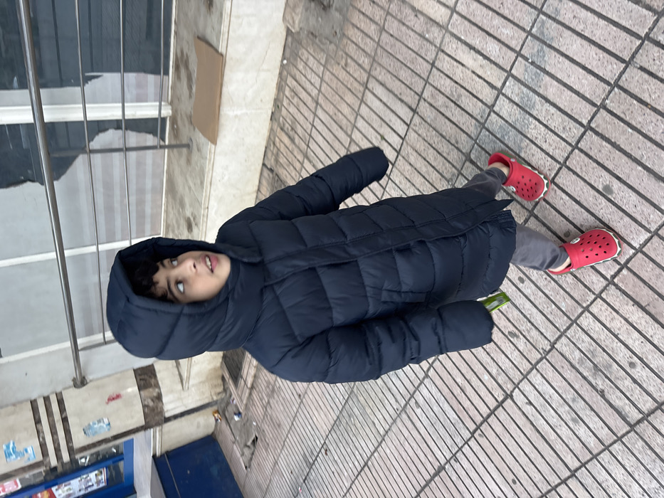 An outdoor, eye-level shot, rotated 90 degrees clockwise, captures a child standing on a paved sidewalk. The child, likely a boy, is positioned slightly off-center towards the left. He has dark hair and is wearing a dark blue (possibly black) hooded puffer jacket that reaches approximately mid-thigh, grey trousers, and bright red Crocs-style shoes with white soles. His head is tilted slightly upwards, and his gaze is directed towards the upper right of the frame, giving him a curious or observant expression. A small, light-colored object, possibly a green or white box or package, is partially visible in his left hand, tucked against his coat.
The background features a tiled sidewalk or pedestrian area made of light-colored, rectangular paving stones laid in a linear pattern. To the left (when upright), there are vertical metal railings, possibly part of a fence or a building's exterior. Behind the railings, reflections suggest large windows or glass panels of a building. Further in the background, a light brown cardboard box lies on the ground. In the lower left corner, there are some faint shapes that appear to be stickers or possibly graffiti on a wall or surface, and a blue shutter or door is visible below them. The lighting suggests it is daytime, and the child's heavy coat implies cooler weather conditions. No discernible text is clearly visible in the image.