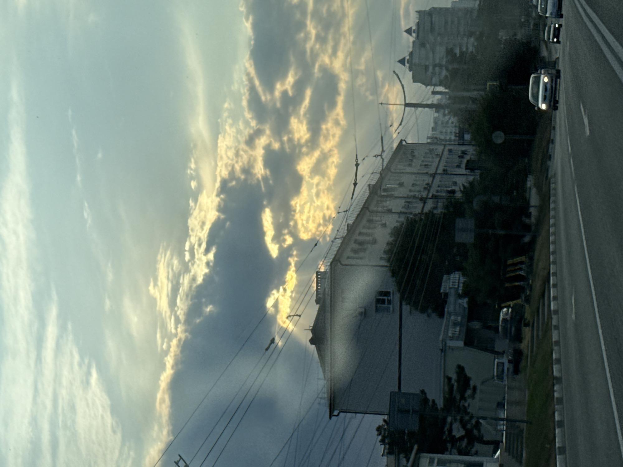 This is a photograph of a street scene in Novorossiysk, Russia, likely taken from a moving vehicle. The sky is filled with dramatic clouds, with the sun breaking through in some areas, creating rays of light. In the foreground, a multi-lane asphalt road is visible, with white lane markings. Several cars are driving on the road, including a silver sedan in the middle lane and a smaller car in the right lane. To the left of the road, there is a sidewalk and a grassy embankment. Behind the embankment, a long, light-colored building with many windows stretches horizontally across the frame. Power lines crisscross the sky above the buildings. Further in the background, other buildings, including some taller, modern structures with distinctive rooflines, are visible. Trees and other vegetation can be seen on the side of the road and around the buildings. The overall impression is of an urban environment bathed in the soft light of either sunrise or sunset.