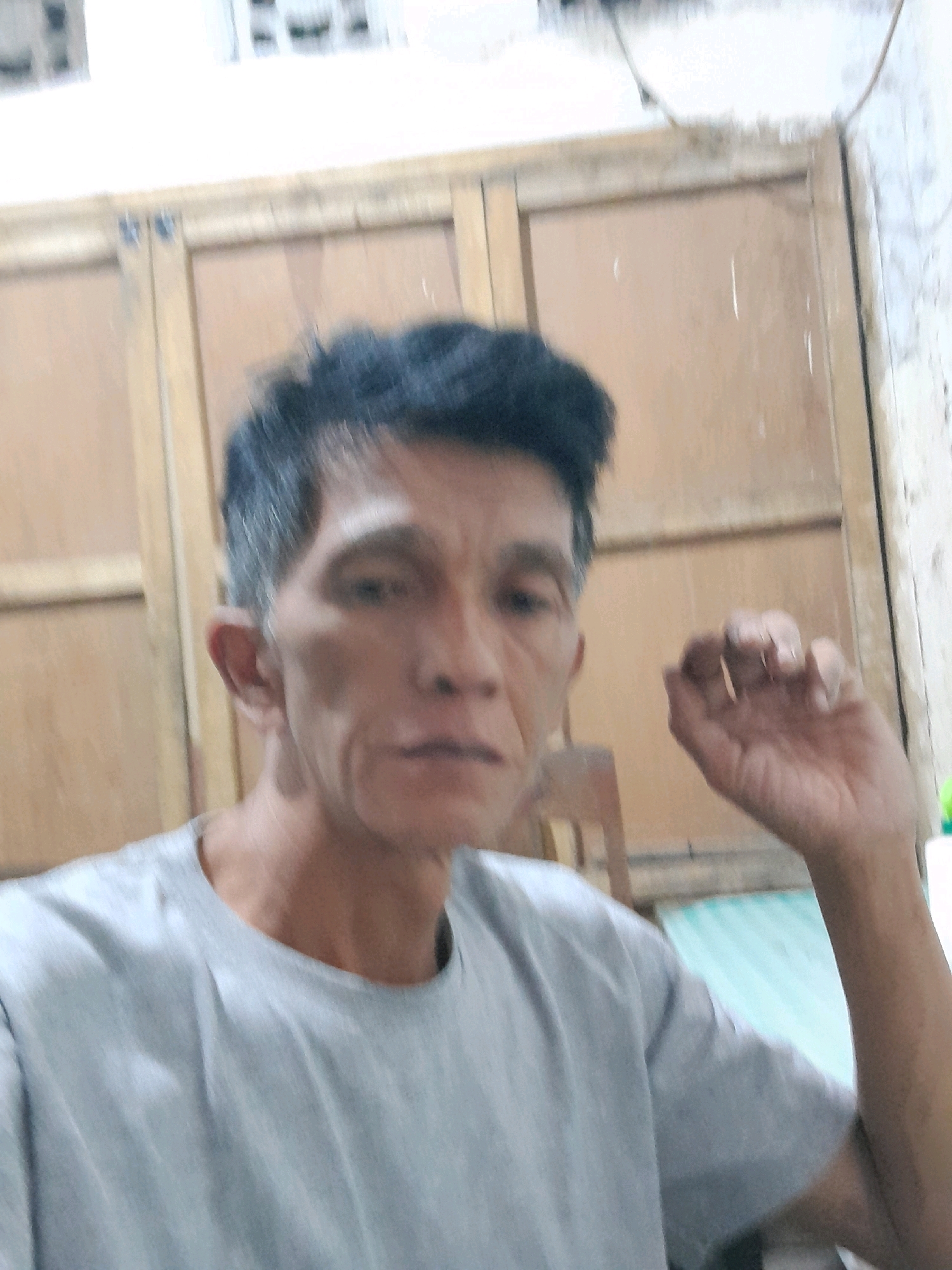 The image shows a medium shot of a man in a light gray t-shirt. He has dark hair with graying at the temples and a lean face with visible wrinkles. His expression is neutral, and his gaze is directed slightly off-camera to his left. His right hand is raised, with fingers slightly curled, near his face. The background appears to be an interior setting, possibly a room with wooden doors or panels. To the left of the man, a wooden chair is partially visible. To the right, a green surface, perhaps a mattress or cushion, is present. The lighting suggests an indoor environment, possibly with ambient light or a diffused light source. The overall scene is casual and appears to be a personal photograph taken indoors. There is no specific information about the location in Glanggang, Indonesia, visible in the image.