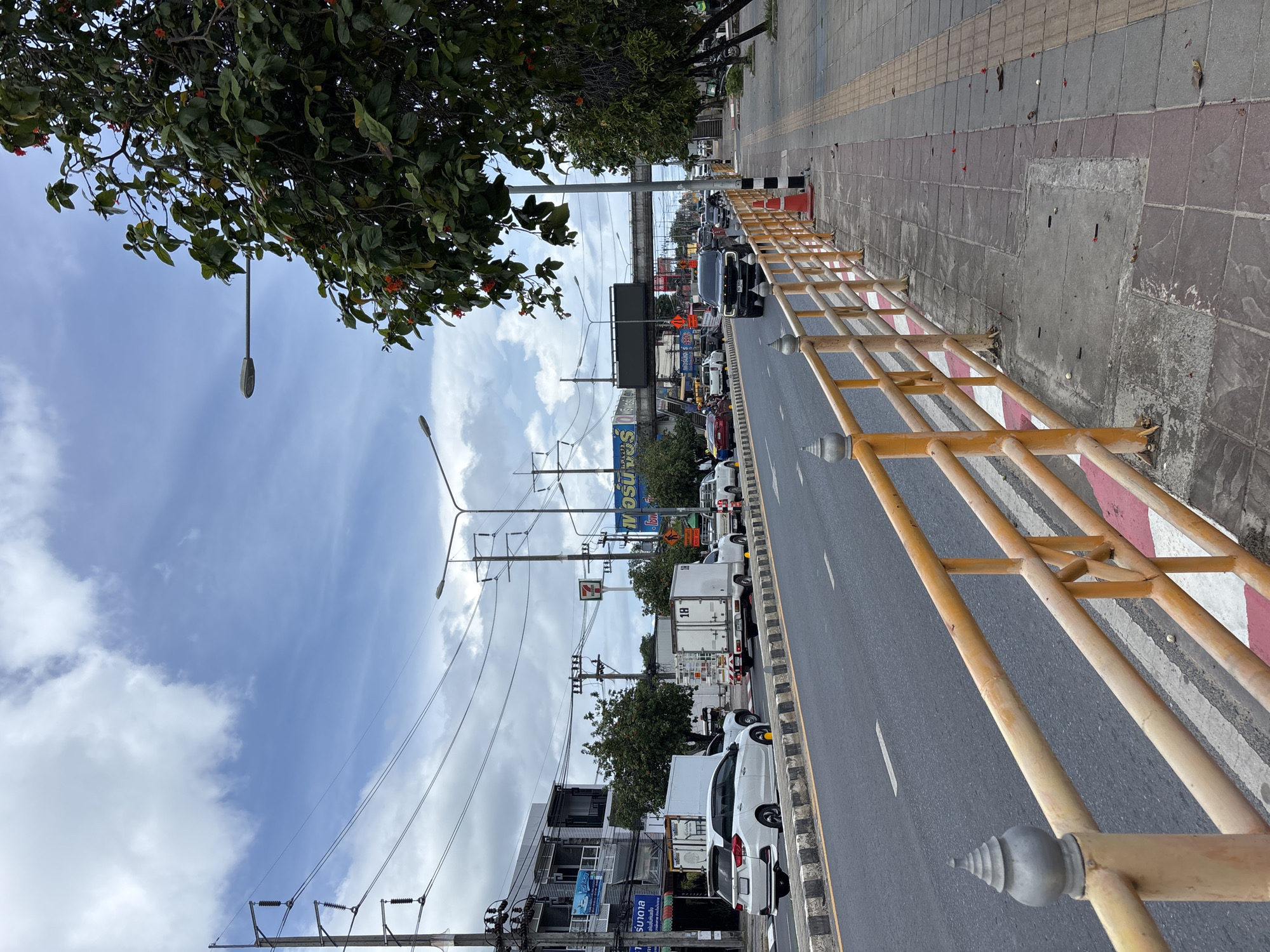 This is a medium-distance, eye-level, exterior shot of a street in Rawai, Thailand. The scene is a busy road with multiple lanes of traffic and a sidewalk lined with a yellow railing. The sky is partly cloudy and the weather appears to be overcast. The road is paved with asphalt and marked with white lines. Several vehicles are visible, including cars, trucks, and motorcycles. The sidewalk is made of paving stones and has a textured surface. There are trees and buildings lining the street, some of which appear to be shops or businesses. Streetlights and utility poles are also visible throughout the scene. The railing in the foreground is yellow with red and white accents and has decorative finials on its posts. The overall impression is of a functional urban environment with active traffic and pedestrian movement.