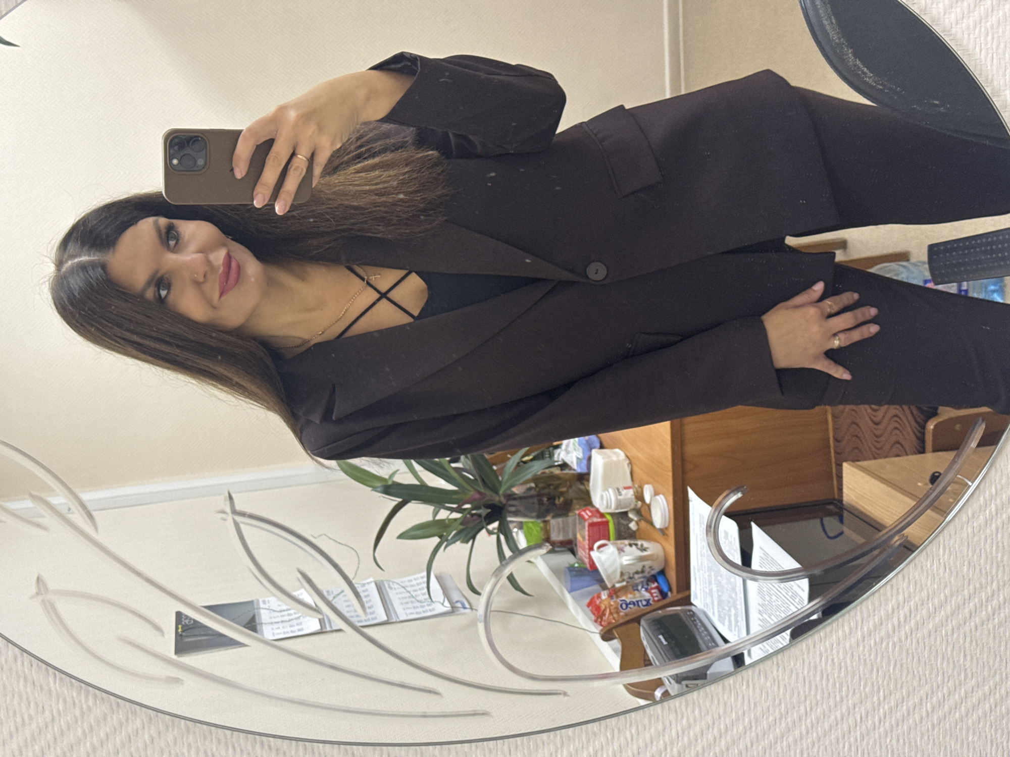 A woman with long brown hair, wearing a dark suit and a black top with a criss-cross neckline, takes a mirror selfie. She is in what appears to be an office or home environment, with a desk visible in the reflection. The desk has various items on it, including a plant in a vase, a mug, snacks, a roll of paper towels, and a small device that could be a radio or speaker. A calendar is also visible hanging on the wall in the reflection. The mirror itself has an ornate decorative edge. The overall impression is of a casual, personal moment captured in an indoor setting.
