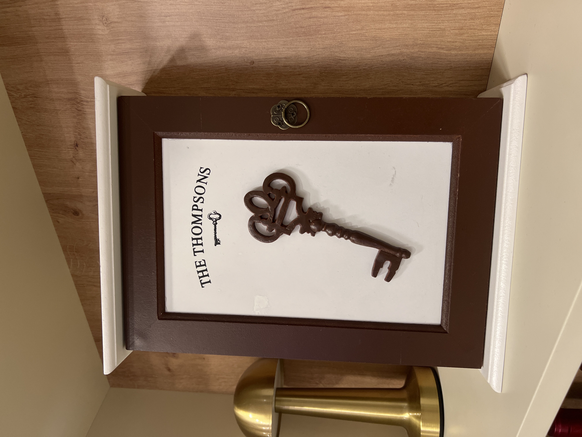 The image shows a decorative object, likely a key holder, placed on a wooden surface. It features a brown frame with a white interior. Inside the frame, the words "THE THOMPSONS" are printed vertically in black, accompanied by a decorative key illustration. A larger, three-dimensional brown key is positioned across the center of the frame. At the top center of the frame, there's a metallic, circular handle. A gold-colored object, possibly a lamp or stand, is partially visible below the key holder. The setting is indoors, on a light-colored wooden or laminate surface. There is no information available from the picture regarding the city of Yerevan or Armenia.