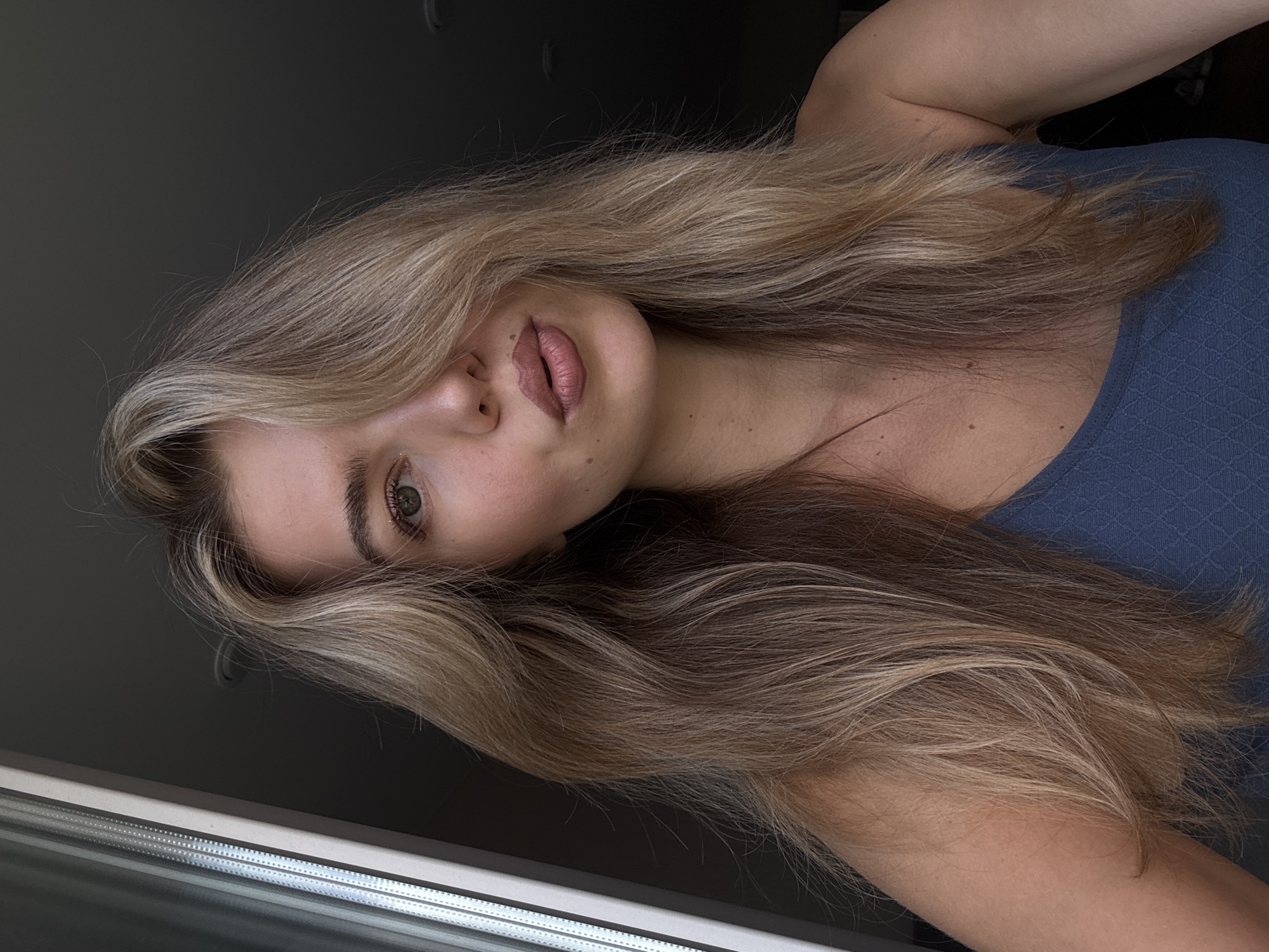 A woman with long, blond hair is indoors, with the lower part of a window visible on the left. She is wearing a blue, textured top. The background is a dark, possibly gray, wall. Her face is angled towards the camera, with her hair partially obscuring one eye.