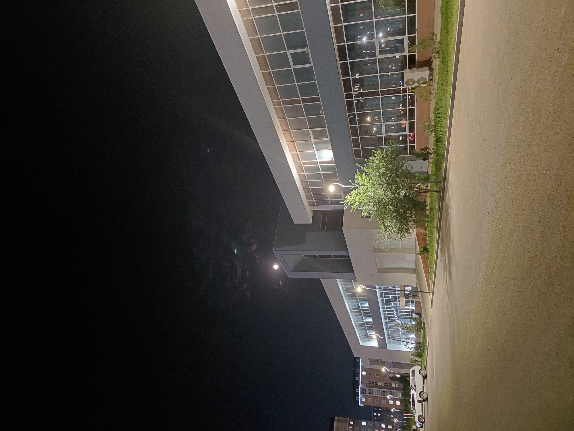 This is a night-time photograph taken outdoors in Aktau, Kazakhstan, showing a modern, multi-story building with a gray and white facade. The building features extensive glass windows that reflect interior lights. In the foreground, there is a paved area, likely a parking lot or plaza, with a concrete curb and a strip of grass and small trees. Two white vehicles are parked on the left side of the paved area. In the background, to the left, are taller apartment buildings with illuminated windows. The sky is dark with some clouds, and the moon is visible. An outdoor air conditioning unit is visible on the ground next to the building on the right.