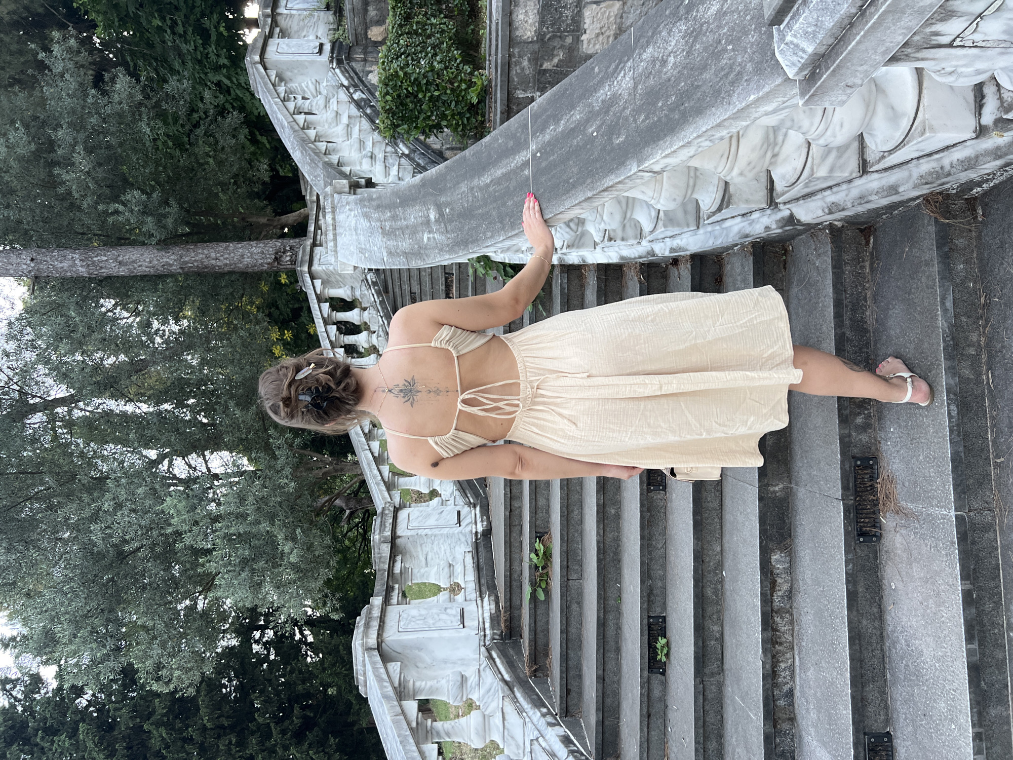 The image shows a woman from behind, walking up a wide, ornate stone staircase outdoors. She is wearing a light-colored, sleeveless dress with a low back and thin straps tied in a bow. Her hair is styled in an updo with a clip. She is holding a small, light-colored purse in her left hand and resting her right hand on the stone railing of the stairs. The staircase is flanked by a decorative marble balustrade with carved details. Green trees and foliage are visible in the background, suggesting a park or garden setting. The steps are made of dark stone and show some signs of wear, with small plants growing in some crevices. The lighting suggests it is daytime, possibly late afternoon. The location is identified as the stairs leading to King Nikola's Palace (now the National Museum of Montenegro) in Podgorica, Montenegro.
