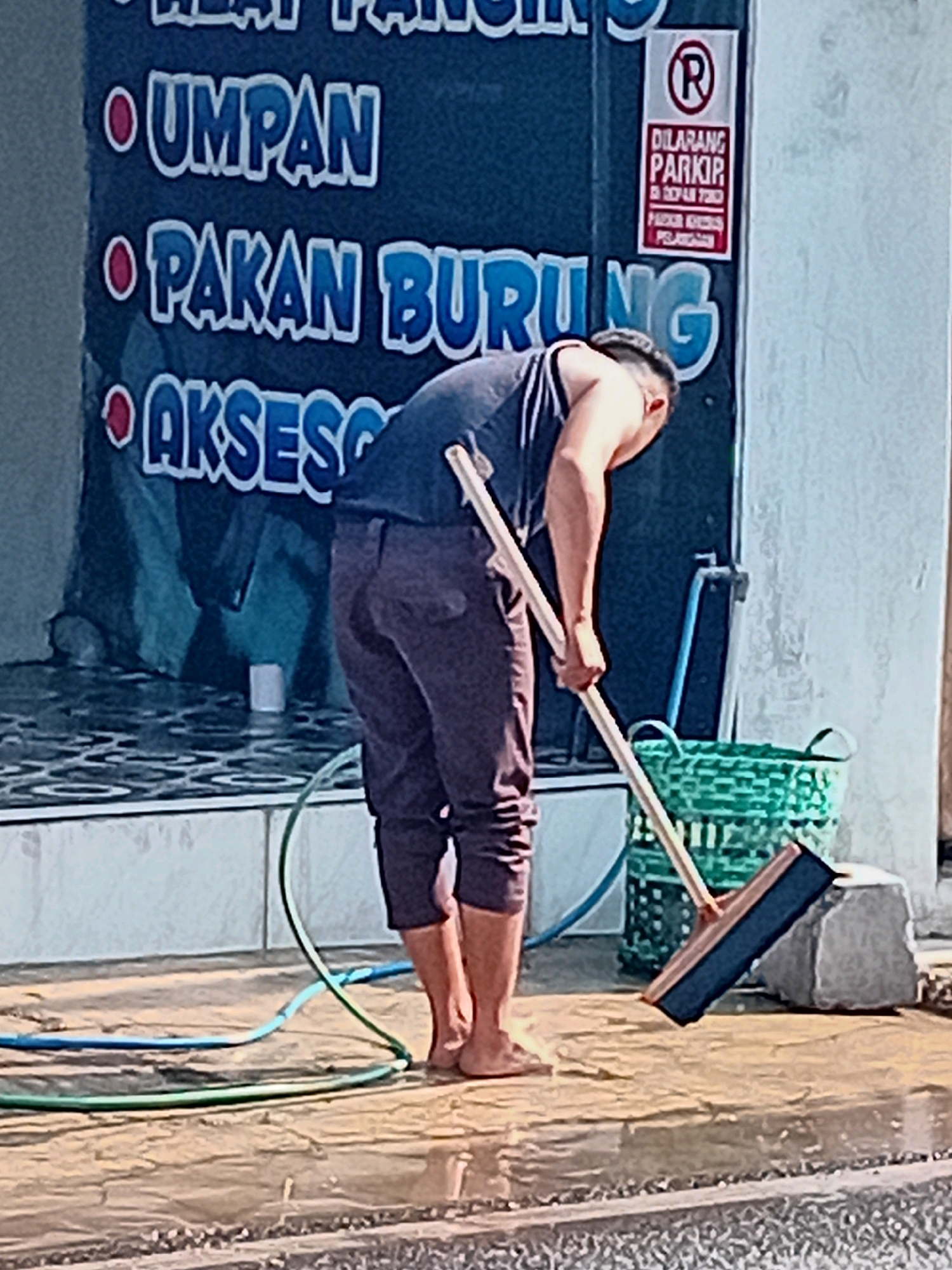 A man is seen from the waist down, wearing dark pants and barefoot, as he sweeps a wet, reflective surface with a wide broom. The broom has a blue head and a light-colored wooden handle. He is positioned to the right of a green woven basket and a light-colored stone. In the background, a dark blue banner with large, white and blue text is visible. The banner includes the words "UMPAN," "PAKAN BURUNG," and "AKSESO," indicating it is likely a shop sign. To the right of the banner, a red and white "No Parking" sign is mounted on a wall. Green and blue hoses are present on the wet ground in front of the man. The overall environment suggests an outdoor commercial area, possibly a shop entrance or walkway.